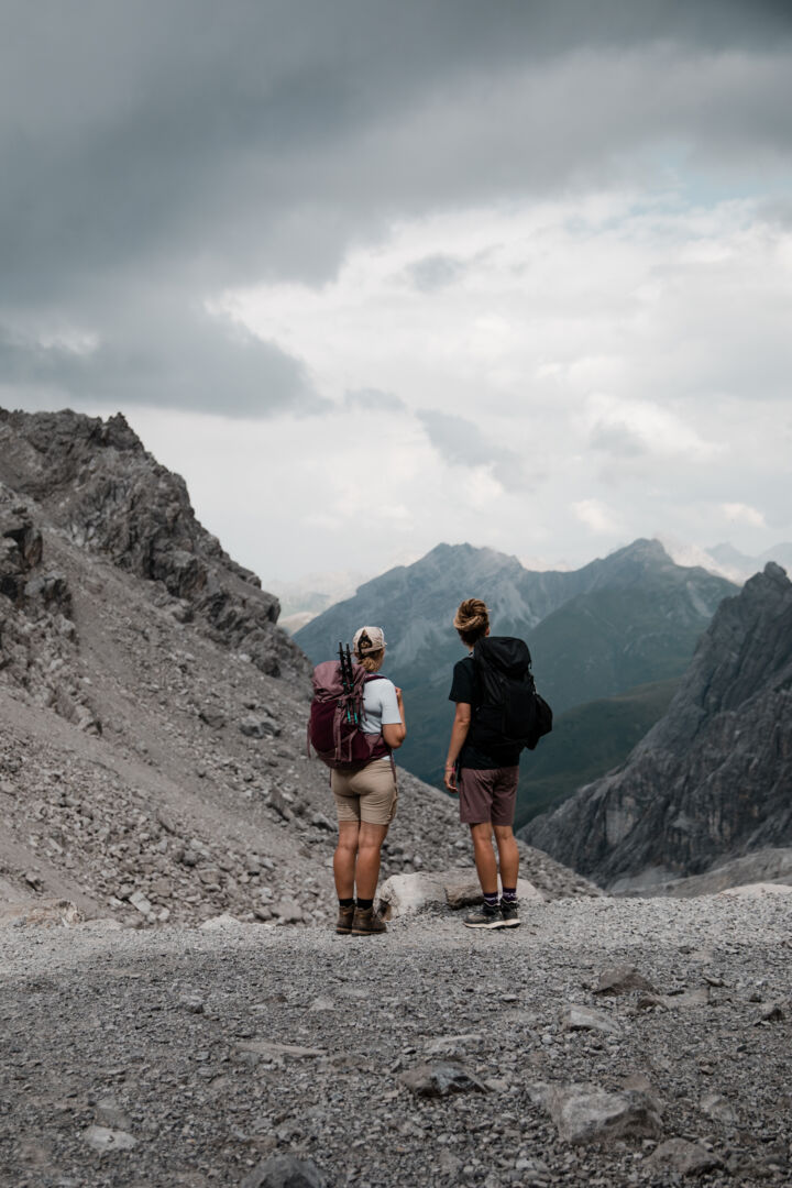 Zwei Wanderer stehen mit dem Rücken zur Kamera auf Geröll und blicken auf ein dramatisches Bergpanorama unter Wolken.