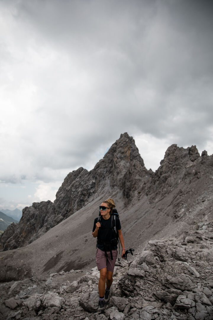 Wanderin mit Rucksack steht auf einem felsigen Grat vor schroffen Gipfeln unter bewölktem Himmel.