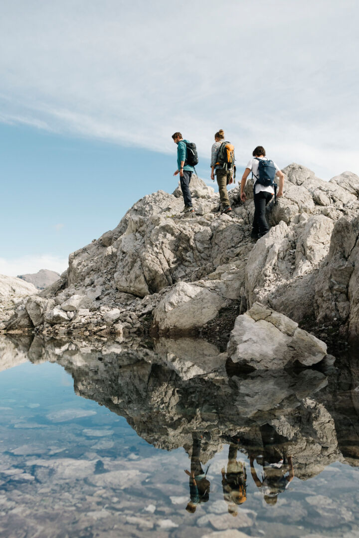 Drei Wanderer gehen entlang eines kleinen Gebirgssees durch karges Gebirge