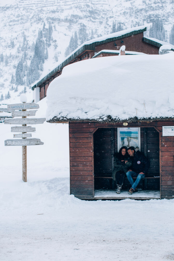 zwei Personen warten in einem holzigen, schneebedeckten Bushäuschen, inmitten der traumhaften Winterlandschaft
