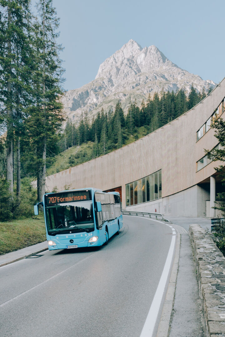Ortsbus fährt auf der Straße und im Hintergrund ist das Biomasseheizwerk von Lech und das Omeshorn