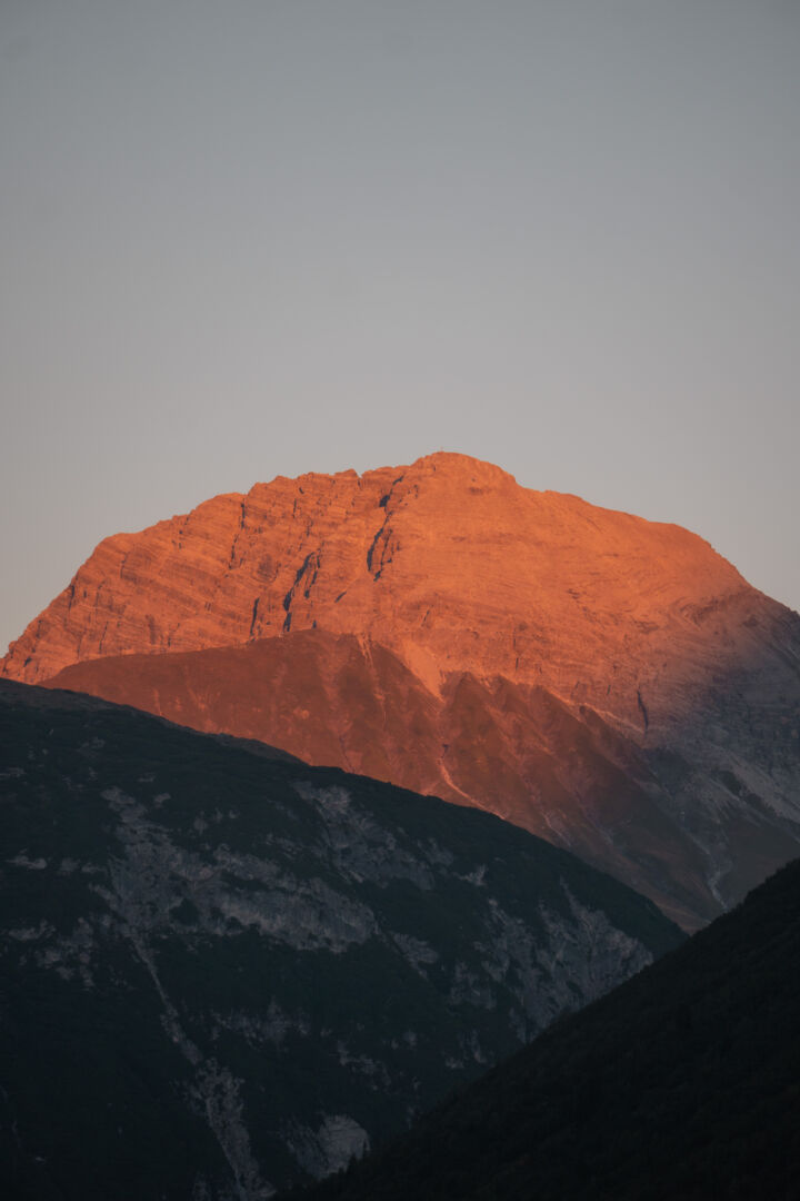 Markanter Berggipfel mit Gipfelkreuz im warmen orangeroten Licht der Abendsonne, davor dunkle, bereits im Schatten liegende Berghänge vor einem grauen Himmel.