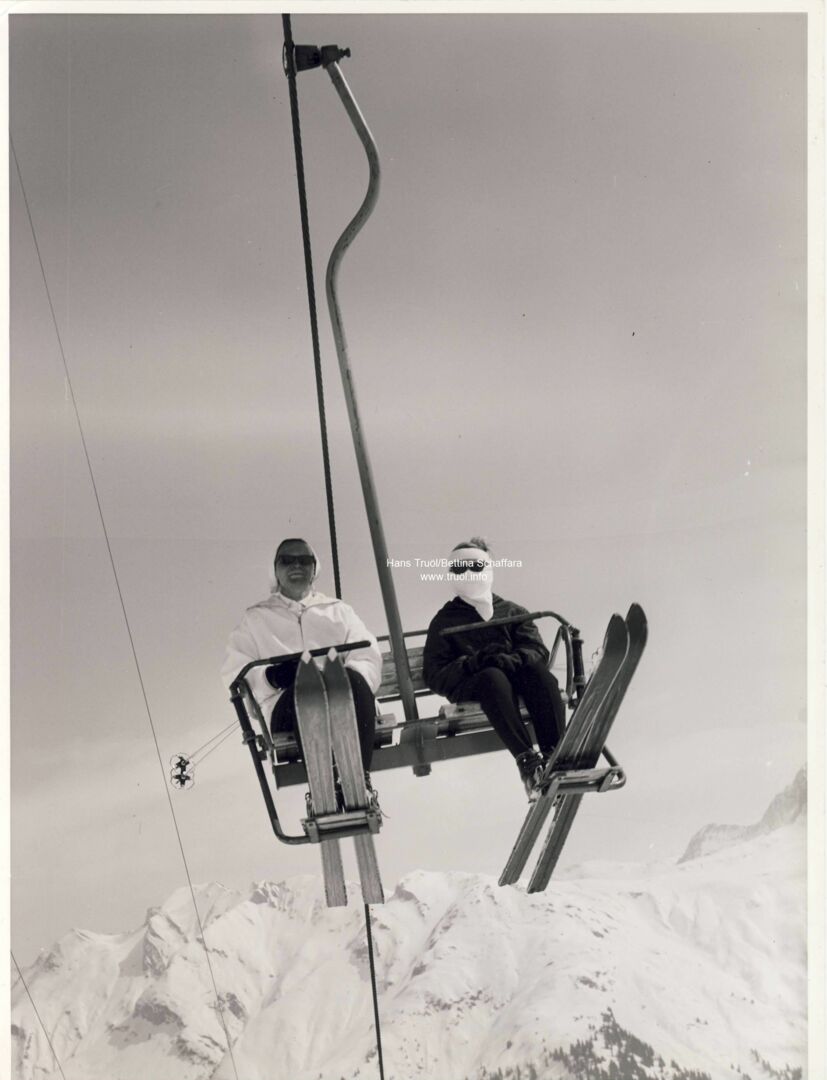zwei Personen sitzen mit ihren Skiern auf einem historischen Sessellift in Lech Zürs, im Hintergrund sieht man die in Schnee gehüllten Bergspitzen