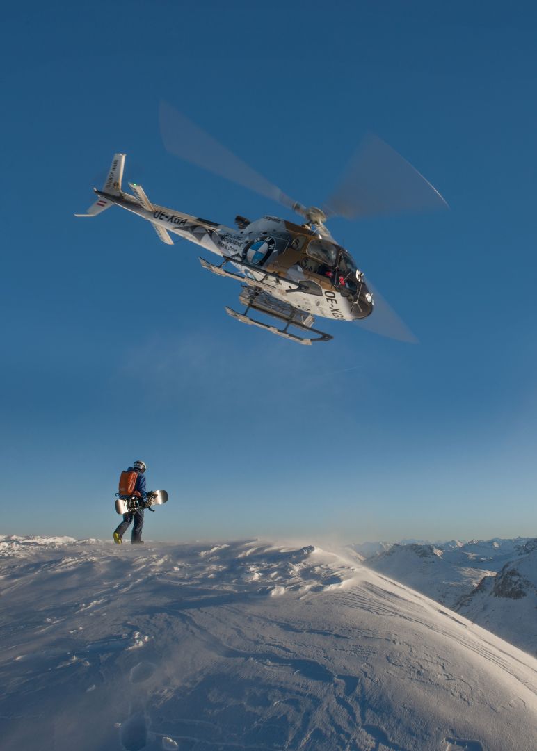 ein Snowboarder steht am Gipfel und betrachtet das malerische Bergpanorama bevor er die unbefahrene Pulverpiste hinunterfährt, während der Hubschrauber über ihm weg fliegt