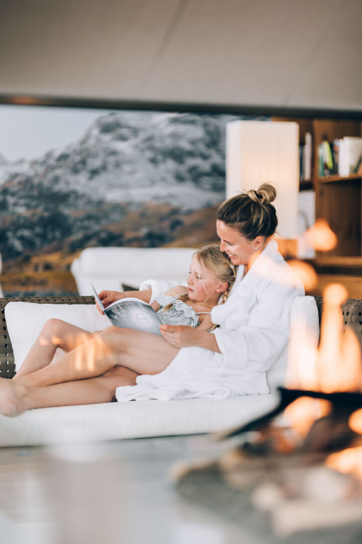 eine Mama liest ihrem Kind eine Geschichte im Wellnessbereich vor im Hintergrund sieht man die atemberaubenden Berggupfel