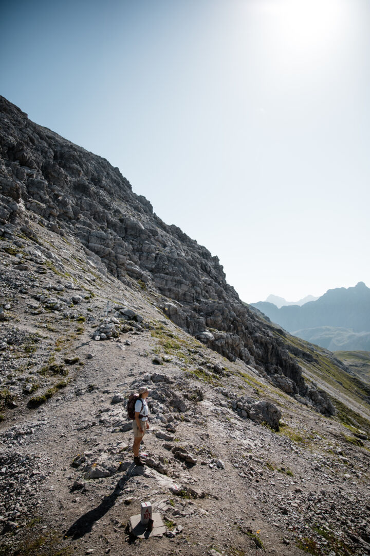 Wanderer steht an einem Wegpunkt mit Steinmarkierung am kargen Berghang, im Hintergrund weitere Gipfel.