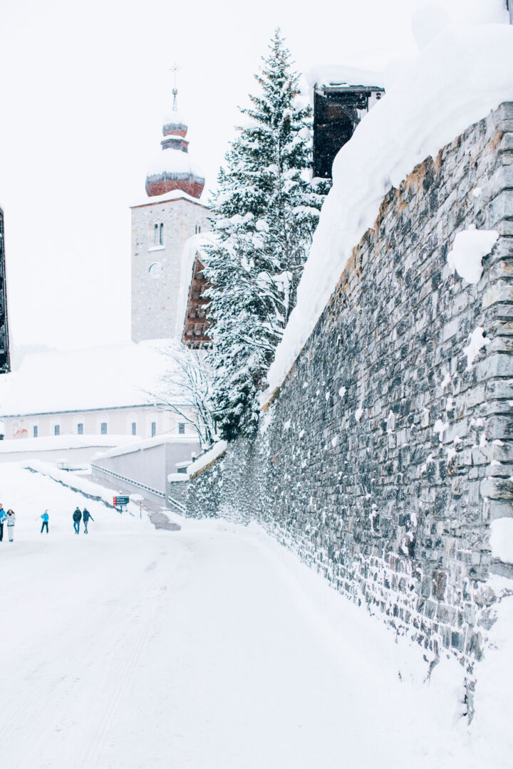 Blick vom schneebedeckten Gehweg auf die Kirche in der Dorfmitte von Lech, davor spazieren einige Personen durch den Schnee