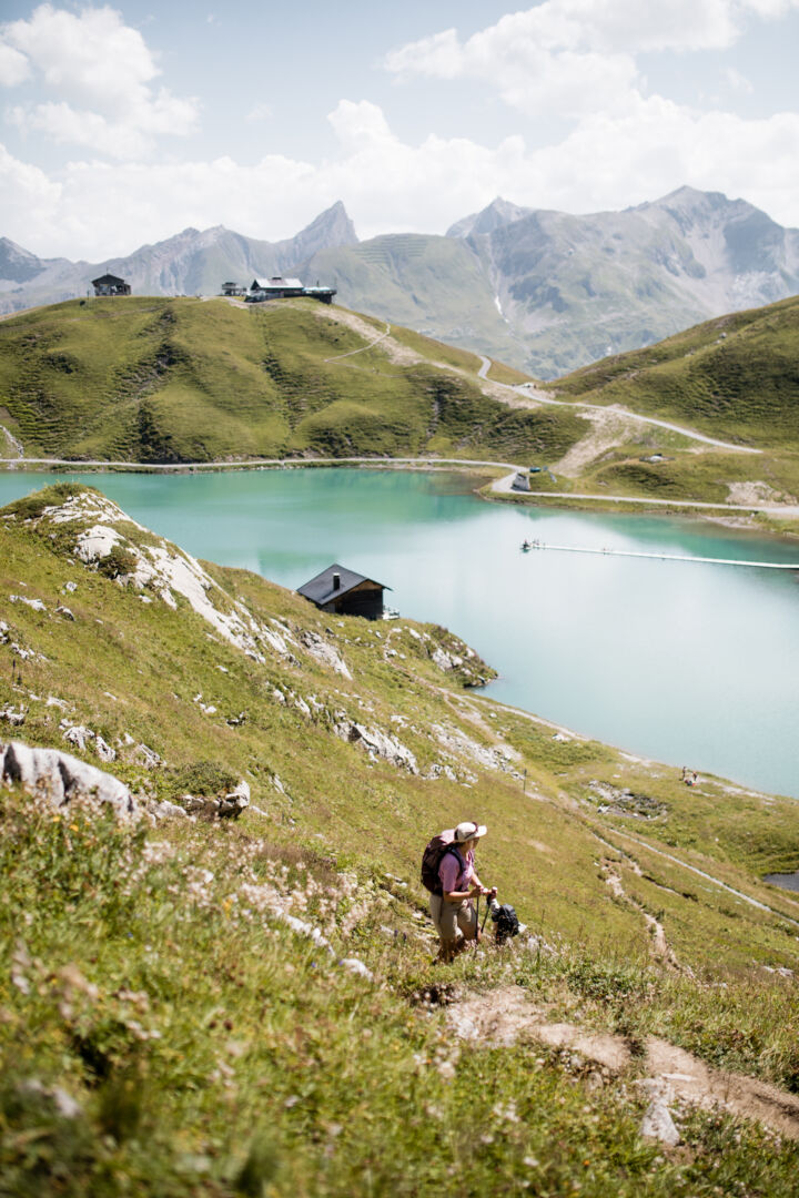 Wanderin mit Trekkingstöcken steigt einen grünen Hang hinauf, im Hintergrund der türkisfarbene Zürsersee.