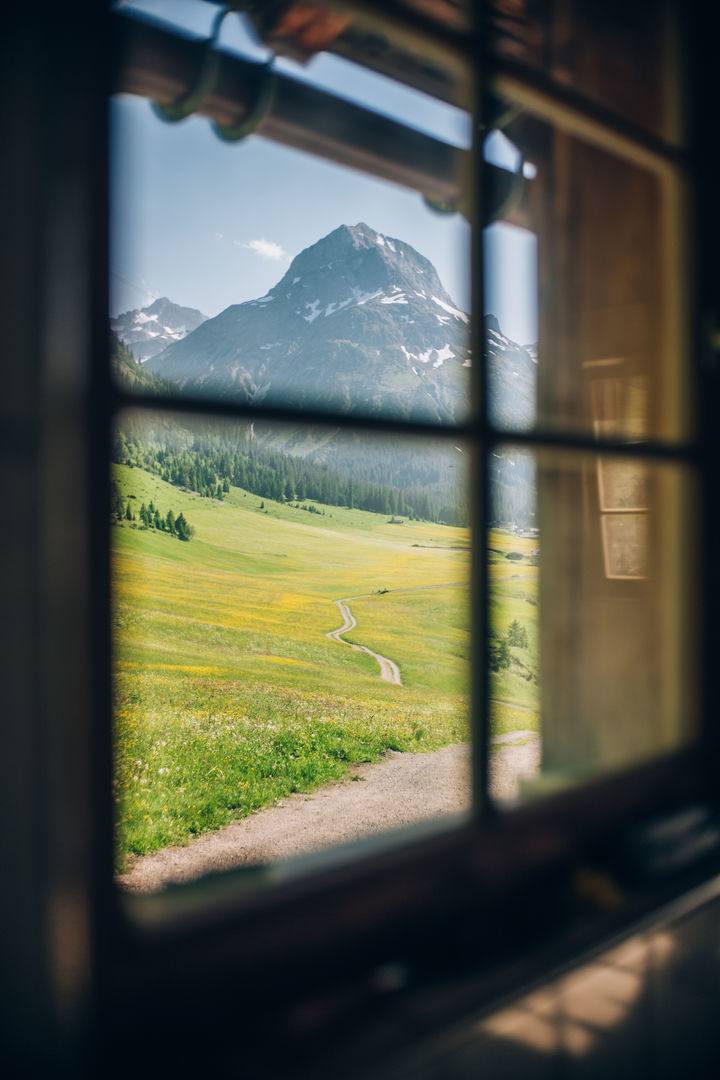 Ausblick aus einem rustikal, traditionellen Fenster mit Blick auf eine blühende Bergwiese und das Omeshorn in der Ferne