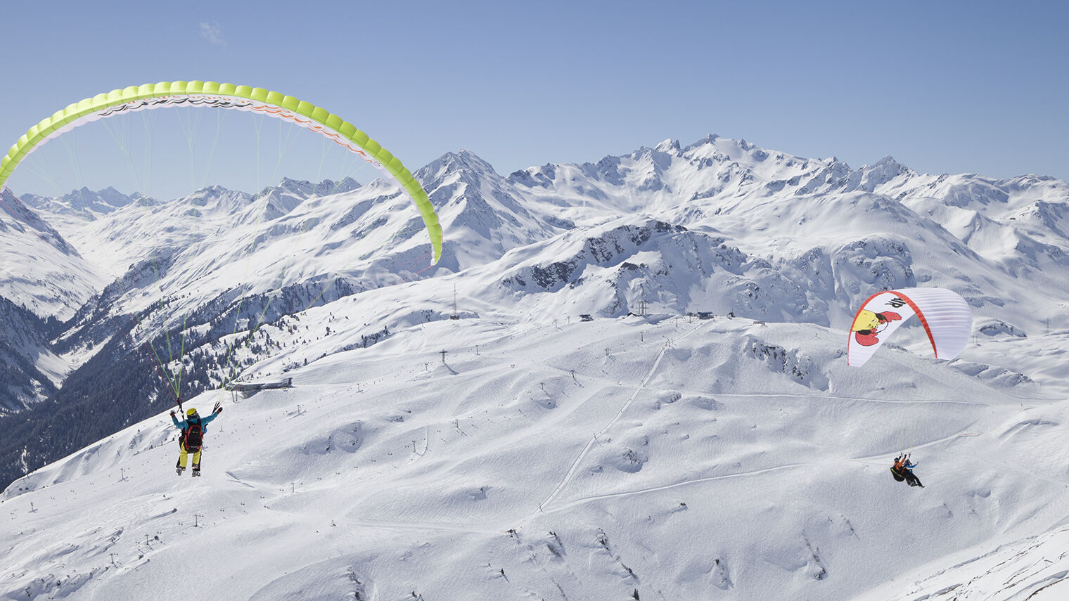 Zwei Paragleiter von hinten zu sehen und im Hintergrund eine große weite zu sehen mit dem Skigebiet und Berggipfeln