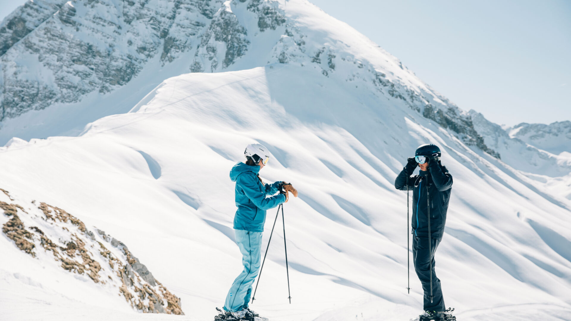 zwei Skifahrer ziehen sich vor der Skiabfahrt an, im Hintergrund sieht man das traumhafte Bergpanorama