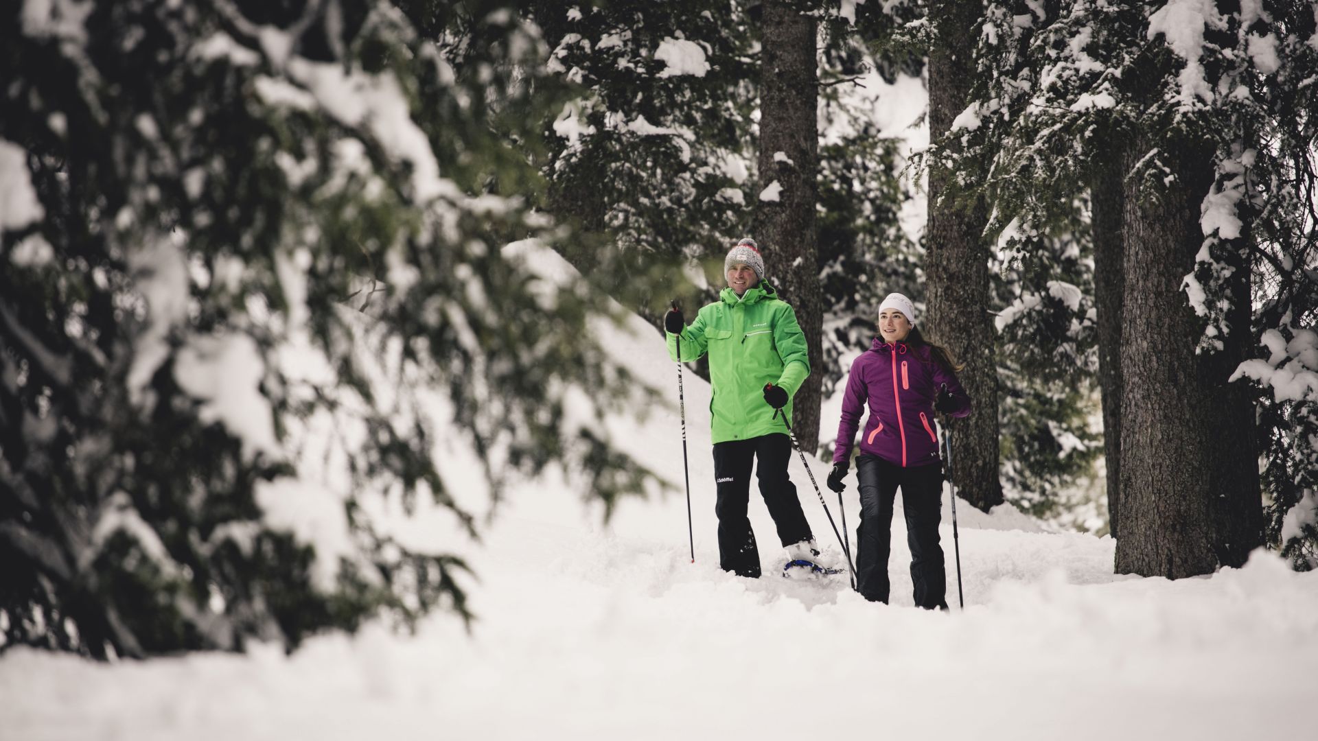 zwei Winterwanderer laufen durch die verschneite Winterlandschaft, im Hintergrund sieht man die schneebedeckten Nadelbäume