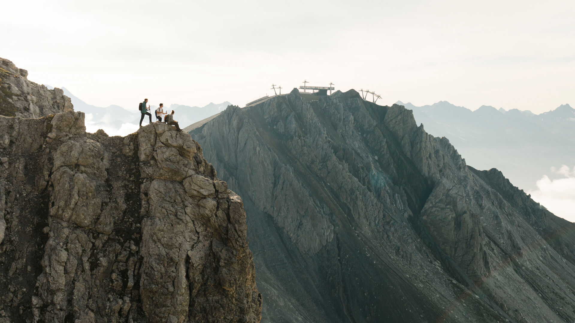 Wanderer sitzen und stehen an einem abschüssigen Felsvorsprung; im Hintergrund befindet sich ein Gebirgshang mit der Bergstation einer Seilbahn