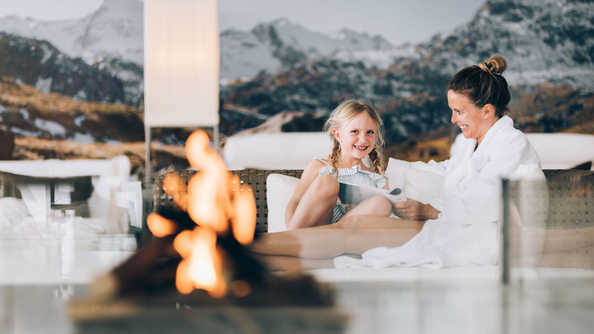 eine Mama und ihre Tochter sitzen gemeinsam im Bademantel im Wellnessbereich, im Vordergrund brennt ein beruhigendes Feuer und im Hintergrund sieht man die verschneite Bergkulisse