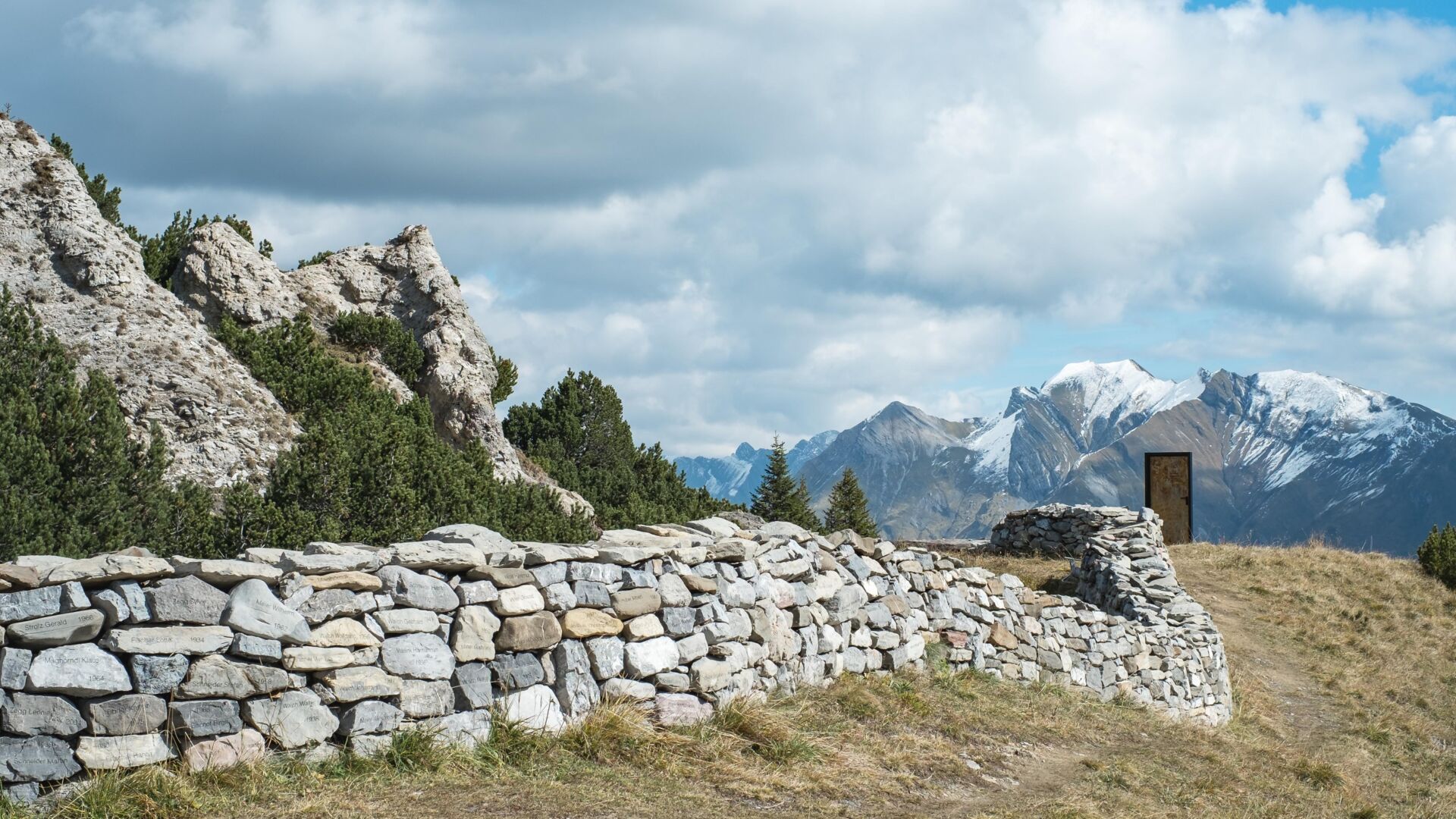 Steinmauer mit einer Türe am Ende der Mauer; im Hintergrund erstreckt sich das Bergpanorama