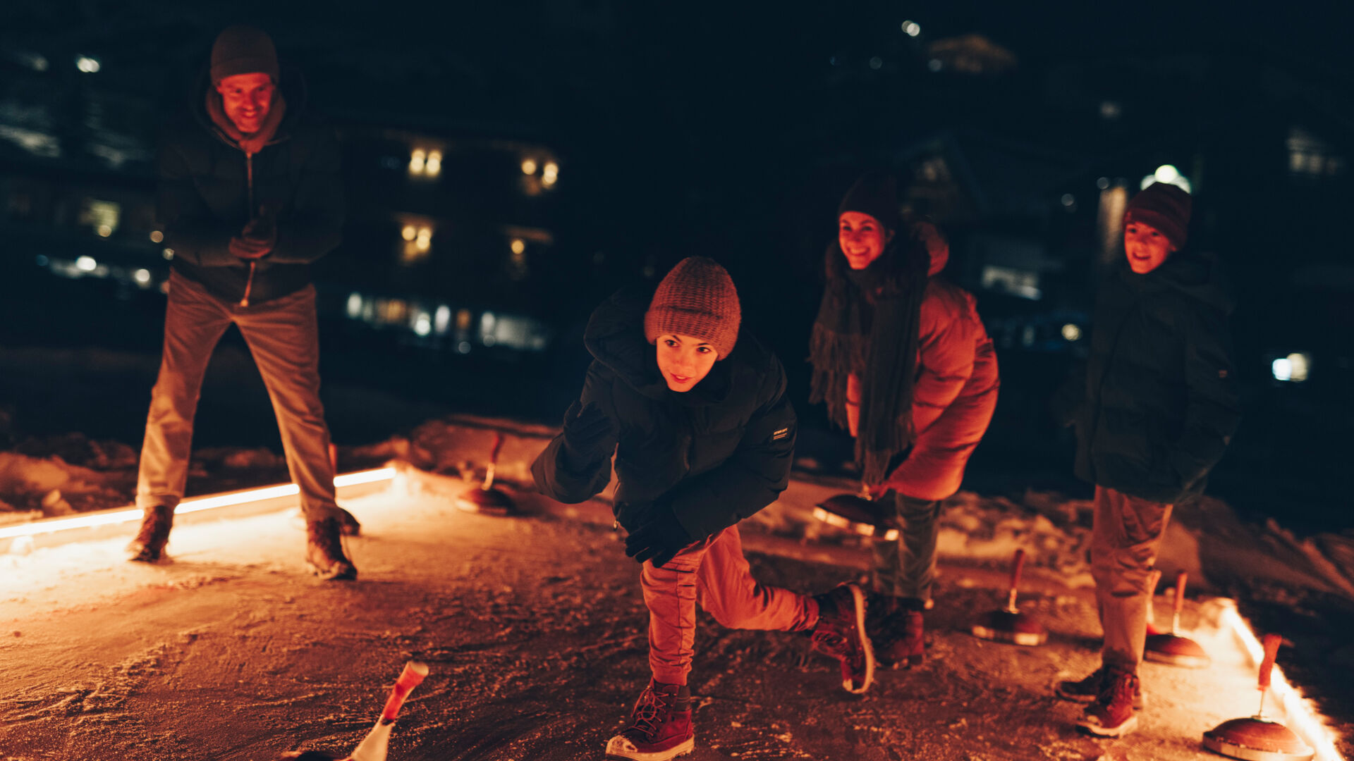 Die gesamte Familie ist auf dem Eisstockbahn und der jüngste Sohn wirft gerade den Eisstock nach vorne auf die Bahn alle schauen gespannt