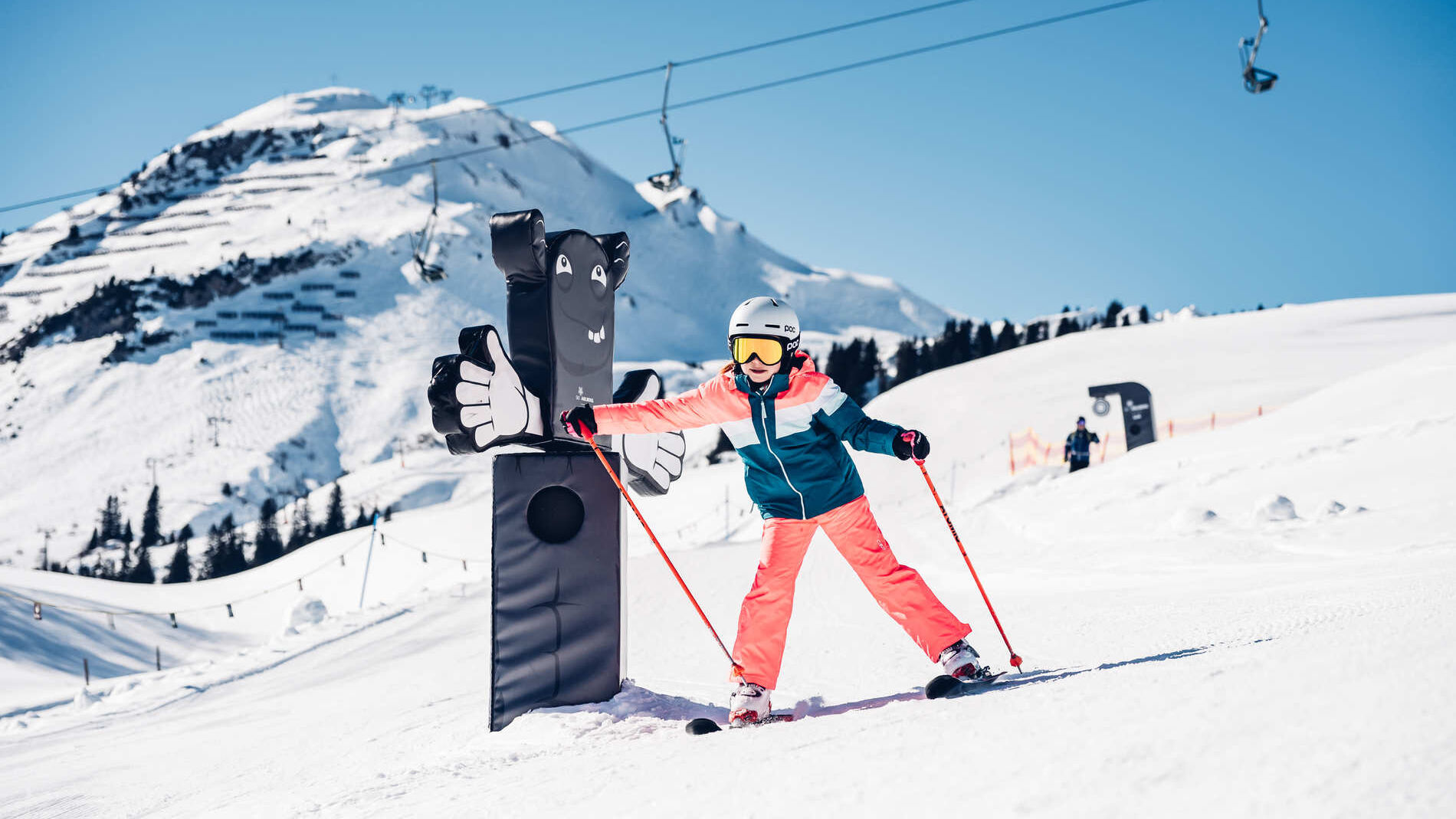 Junge Skifahrerin schlägt lachend gegen eine Daumen-hoch-Figur an einer Spielstation im Skigebiet Lech, umgeben von Bergpanorama und Sessellift.