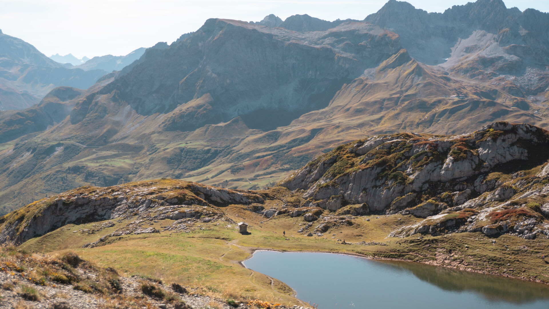 Blick auf einen kleinen Bergsee mit einer Steinhütte am Ufer, umgeben von herbstlich gefärbten Almwiesen und Felsen, im Hintergrund mehrere dunstige Bergketten.