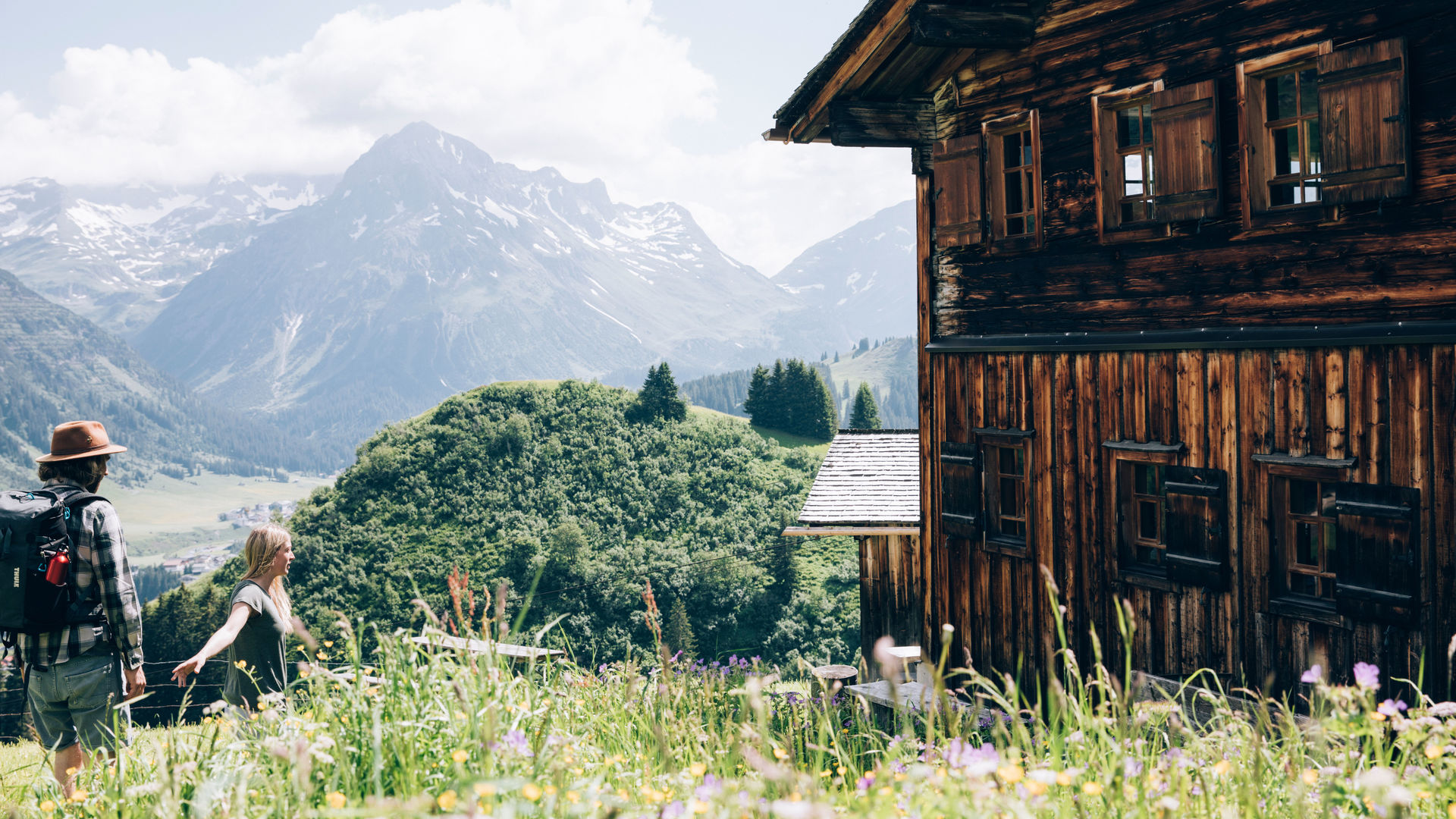 zwei Wanderer laufen durch die blühende Bergwiese an einem urigen Holzhaus in Bürstegg vorbei, mit einem traumhaften Bergpanorama im Hintergrund