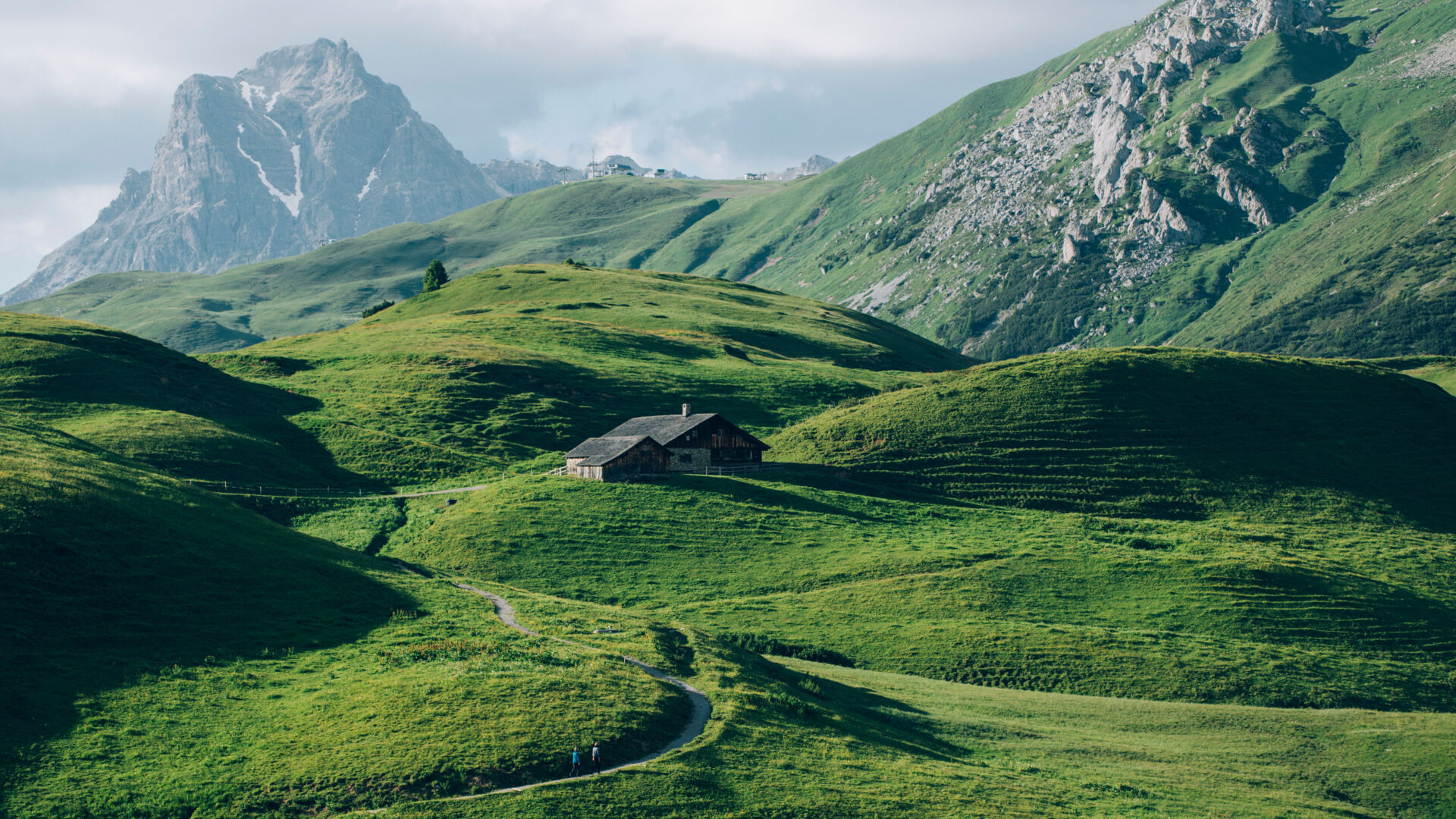 weite grüne Wiesen mit steinigen Berggipfeln im Hintergrund, in der Mitte steht ein Holzhaus