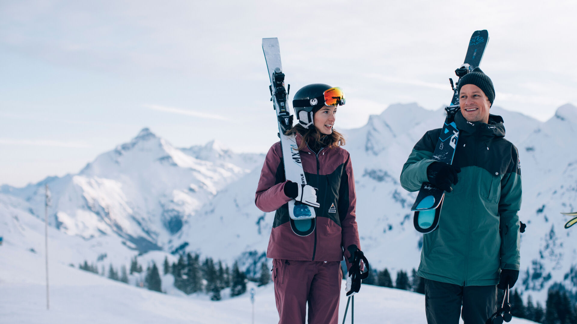 eine Skifahrerin und ein Skifahrer laufen gemeinsam, mit ihren Ski auf den Schultern, zur Skipiste, im Hintergrund sieht man das schneebedeckte Bergpanorama
