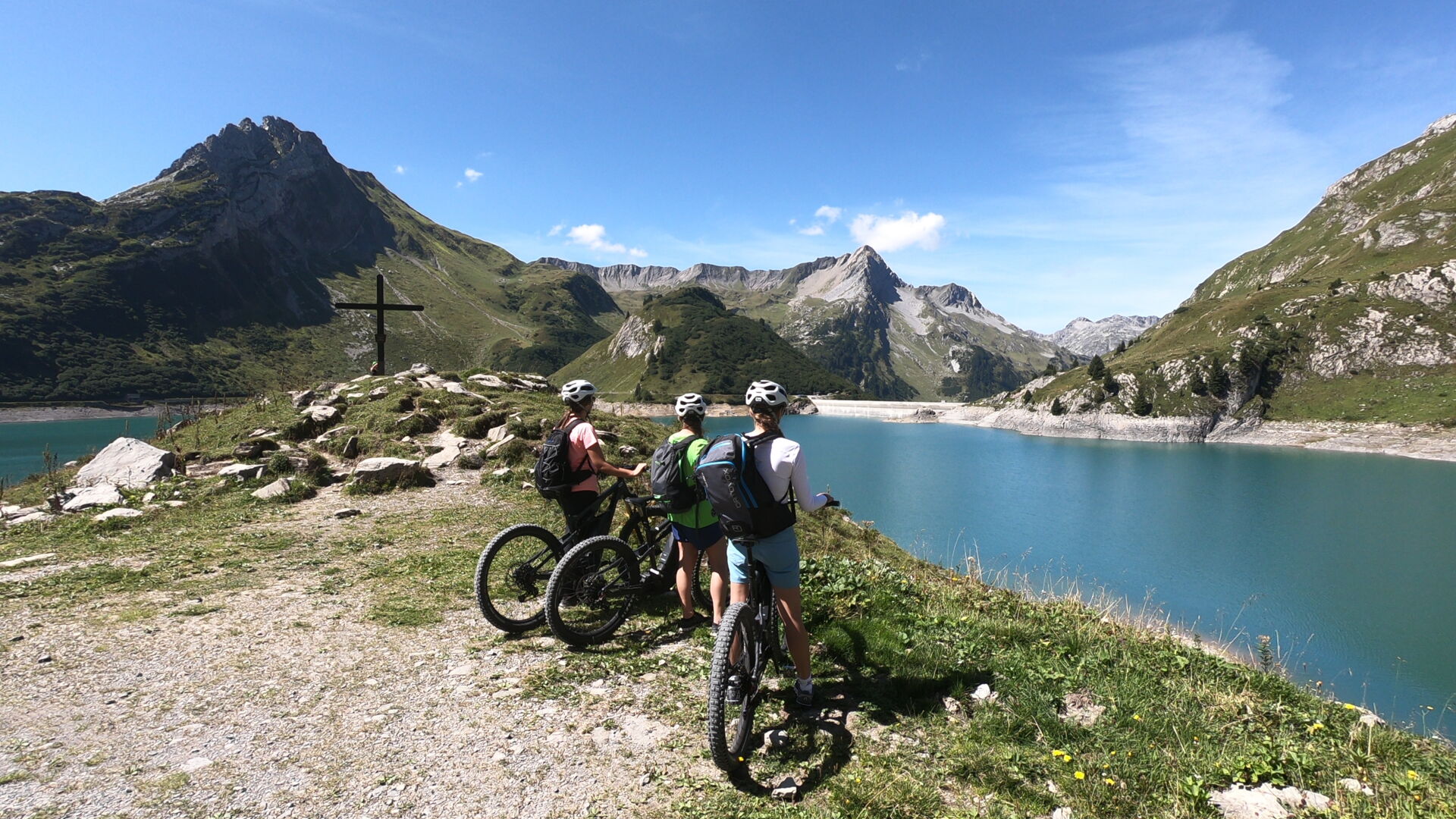 Drei Radfahrer stehen an einem Aussichtspunkt und blicken vorbei an einem Gipfelkreuz über einen türkiesen See auf ein wunderschönes Bergpanorama. 
