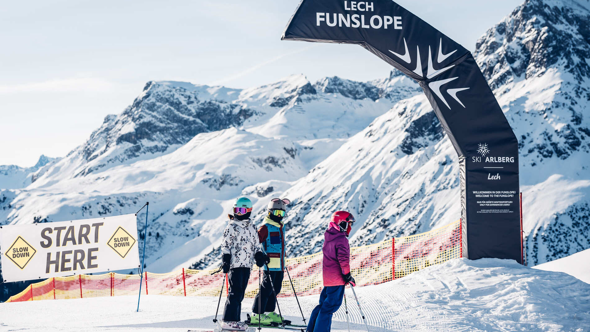 Drei Skifahrer stehen am Startbogen der Lech Funslope, umgeben von schneebedeckten Bergen.