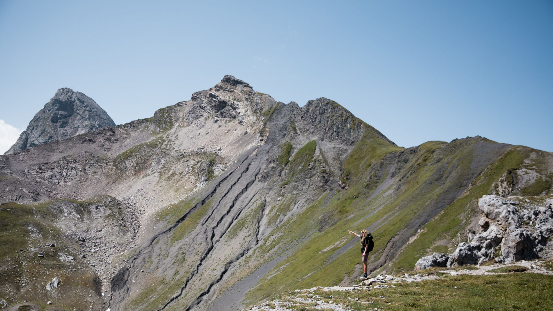 Wanderin zeigt auf einen fernen Gipfel, während sie auf einem grünen Grat mit Blick auf schroffe Berge steht.