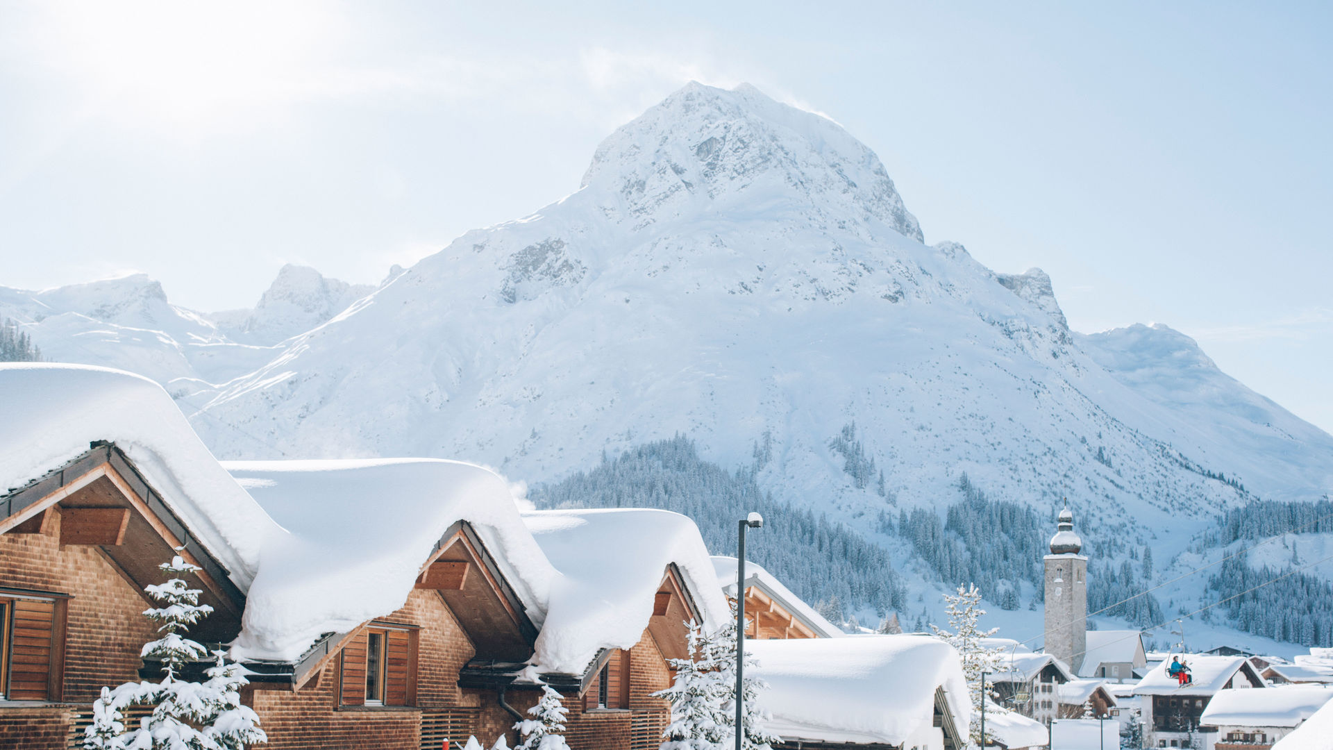 mehrere schneebedeckte Holzhäuser stehen vor der traumhaften Winterkulisse, im Hintergrund sieht man Lech und das Omeshorn