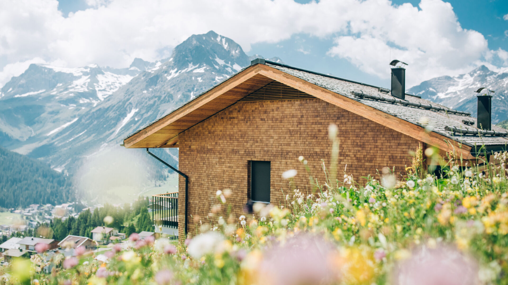 Bunte Frühlingswiese mit einem Holzhaus und einem steinigen Bergpanorama im Hintergrund