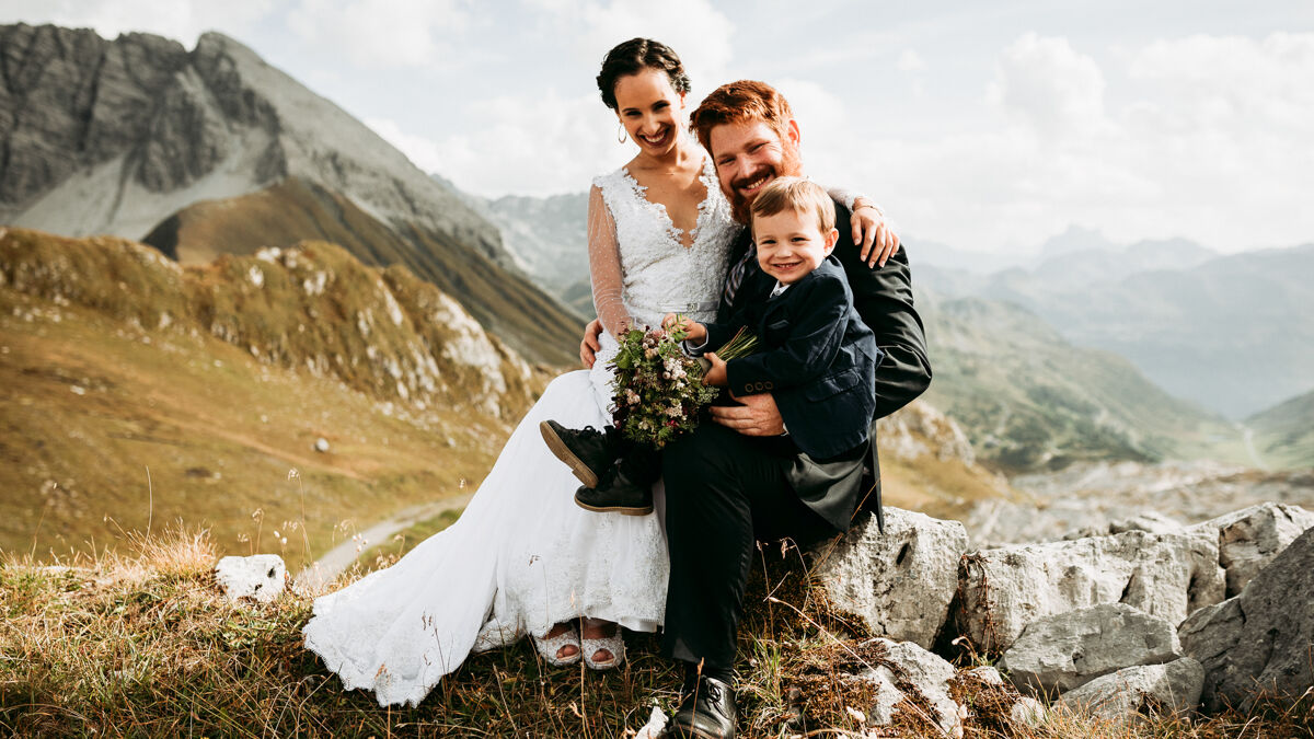 Ehepaar mit Sohn sitzt gemeinsam in Berglandschaft und lächelt in die Kamera