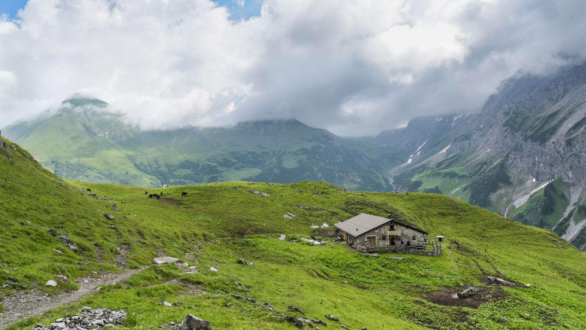Ausblick auf eine Alpe, eingebettet in der Berglandschaft mit grünen Wiesen und Quellwolken am Himmel
