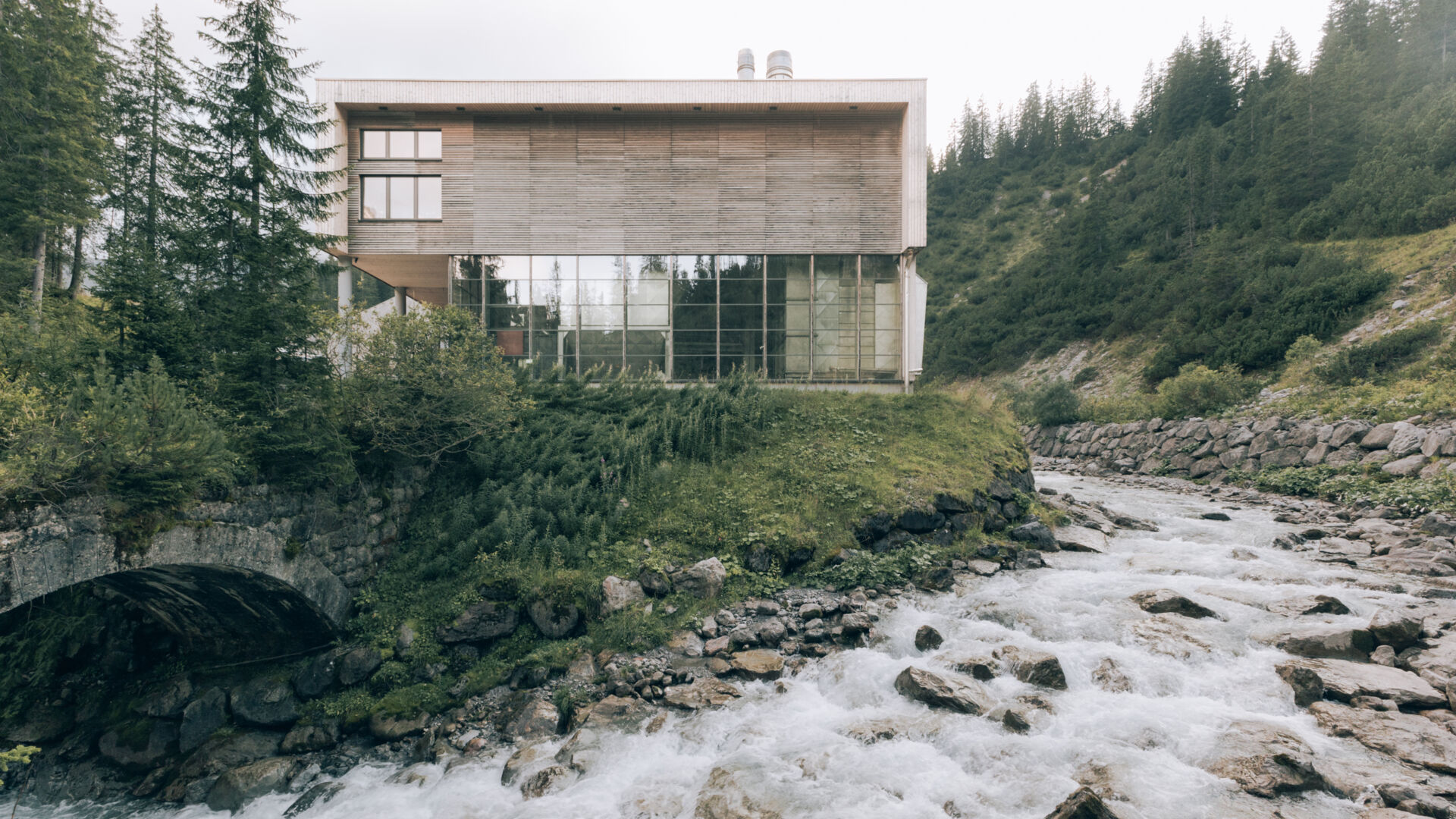 Biomasseheizkraftwerk Zürs ist ein Holzgebäude von der Hinteransicht aus Holz und Glasfassade und nebenan sieht man den Bach mit Wasser