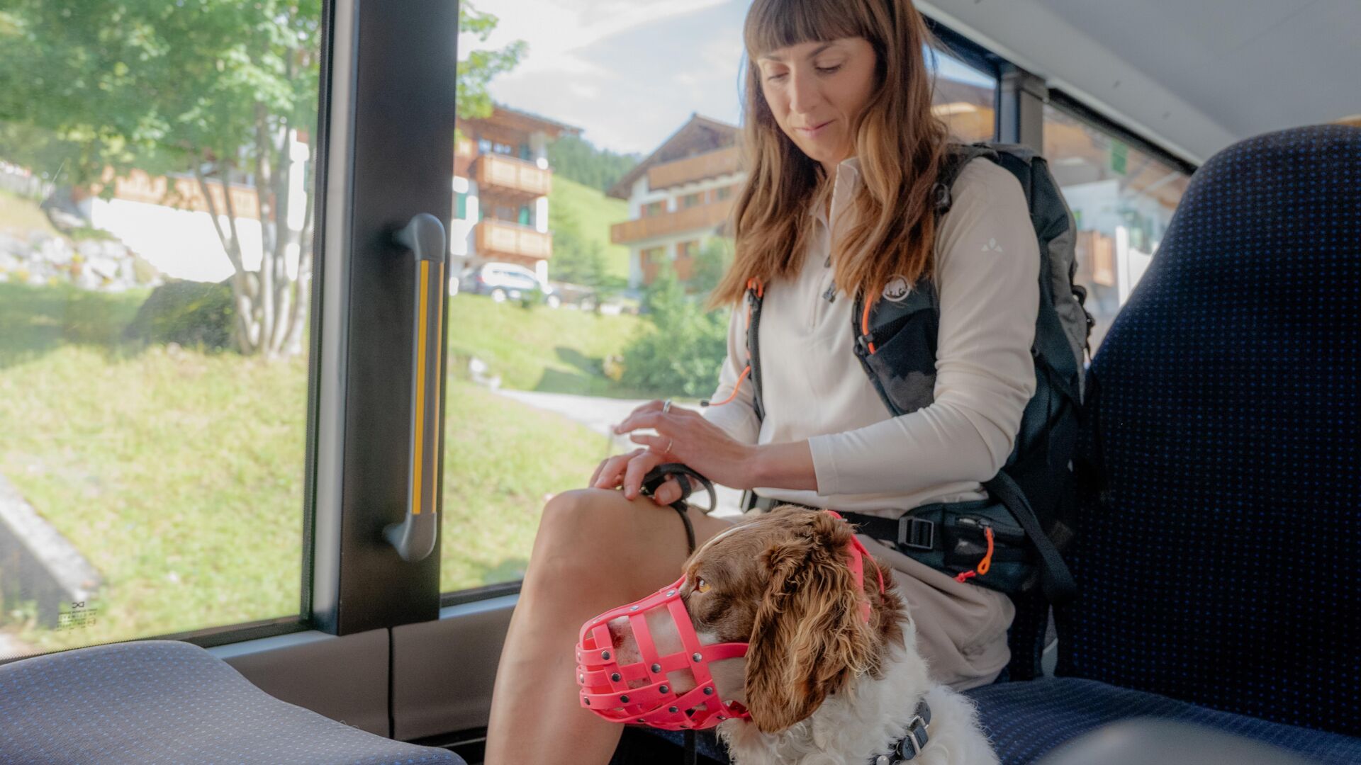 Frau sitzt im Bus und hält die Leine ihres Hundes mit pinkem Maulkorb neben sich.