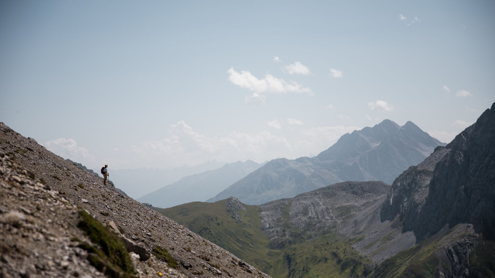 Einzelner Wanderer steht klein auf einem steinigen Hang, im Hintergrund weite Bergketten im Dunst.