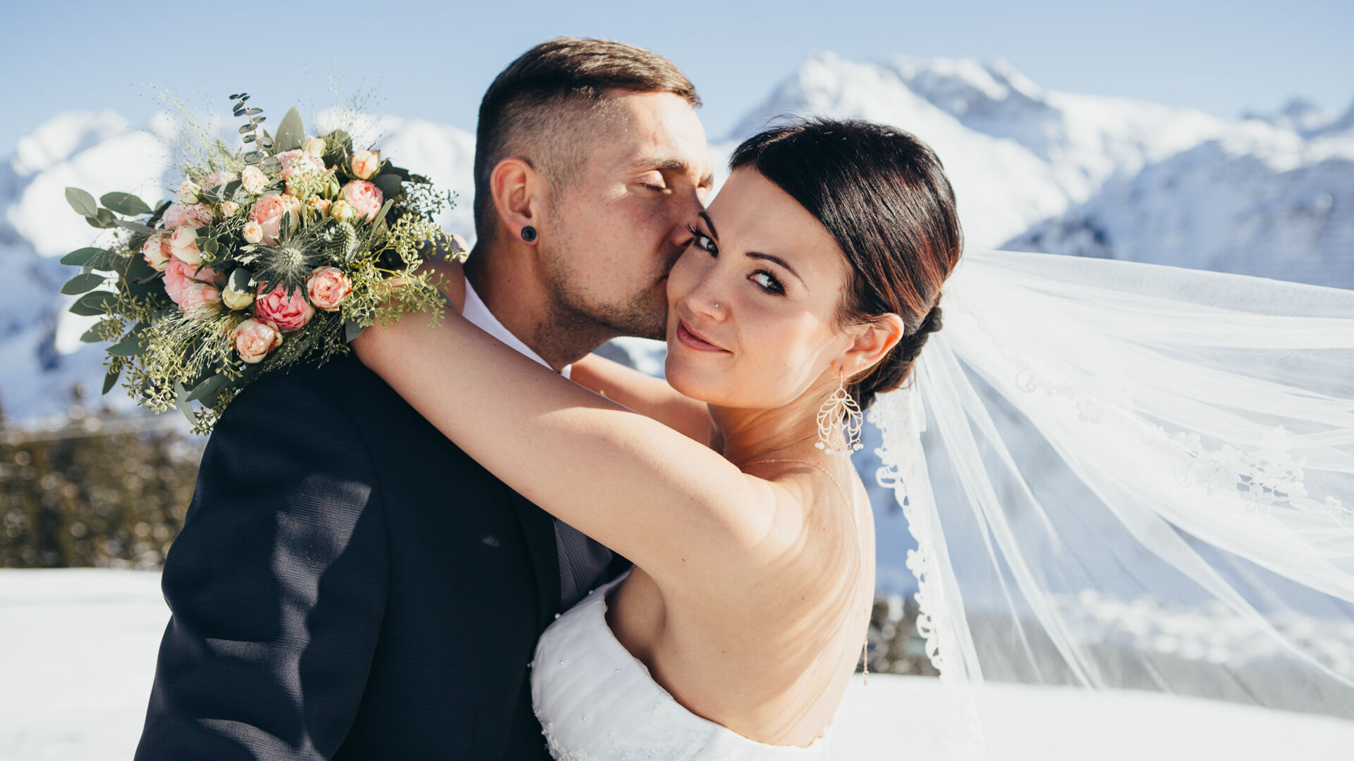 Bräutigam küsst seine Braut auf die Wange, im Hintergrund ragen die verschneiten Berge hoch
