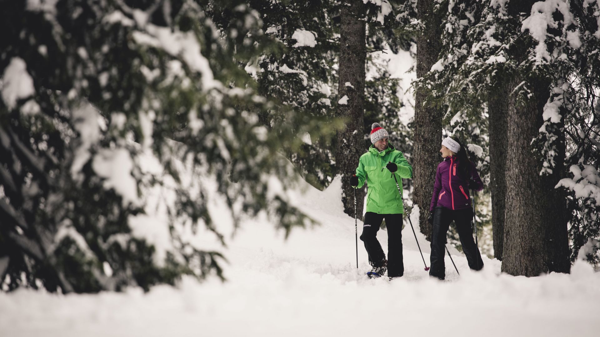 zwei Personen spazieren gemütlich über den Winterwanderweg, im Hintergrund sieht man die verschneiten Nadelbäume