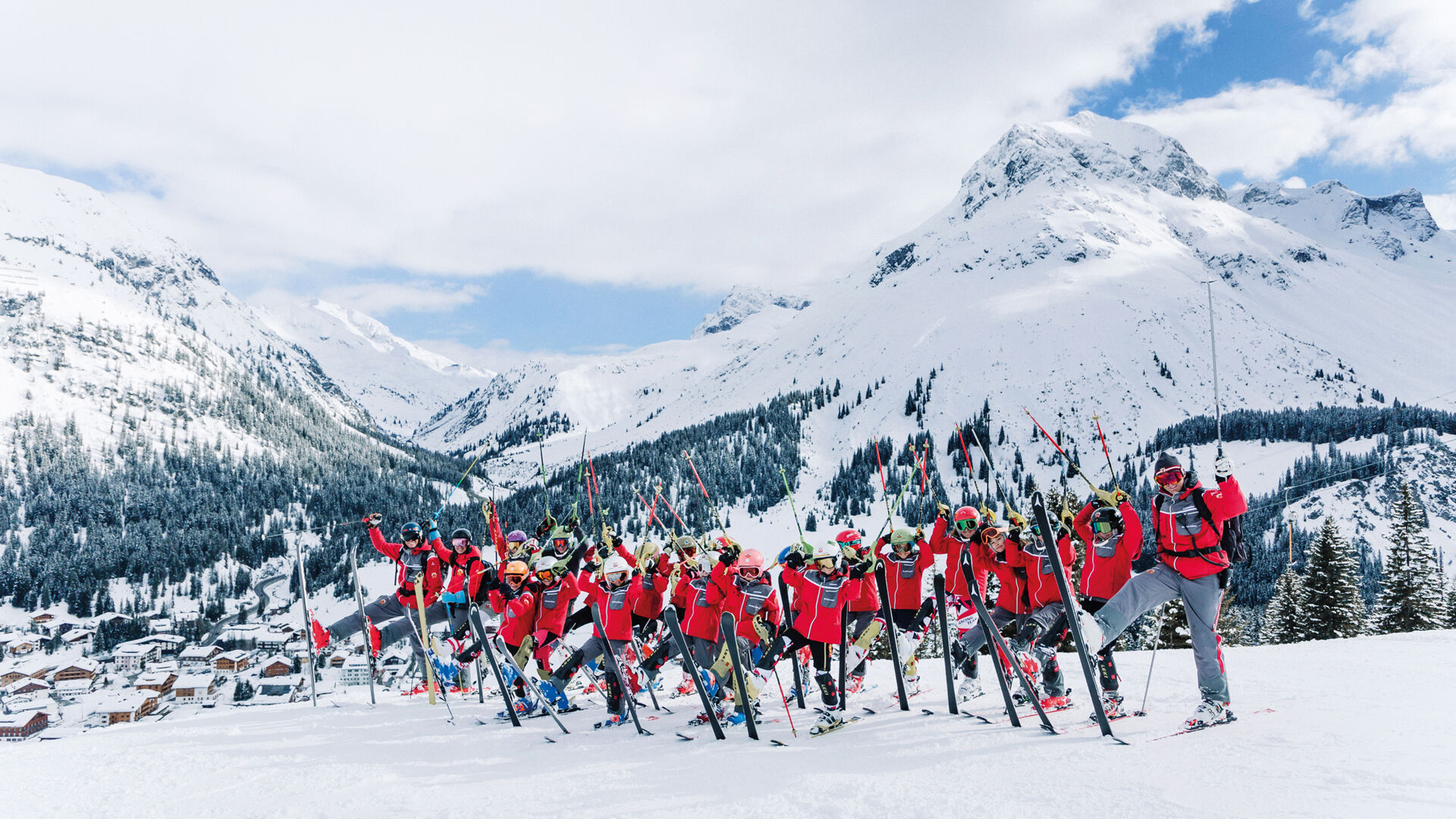 der Skiclub Lech posiert auf der Skipiste mit einem aufgestellten Ski vor dem winterlichen Bergpanorama 