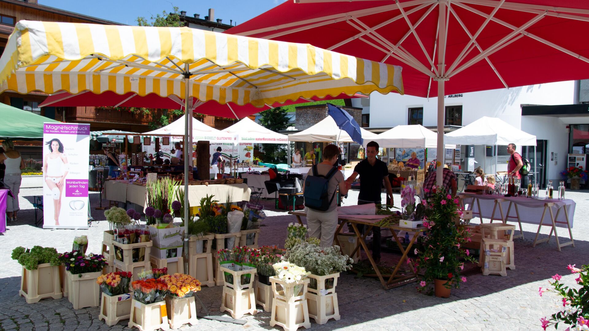 Tannbergermarkt im Freien mit bunten Blumen, Verkaufsständen und großen Sonnenschirmen an einem sonnigen Sommertag.