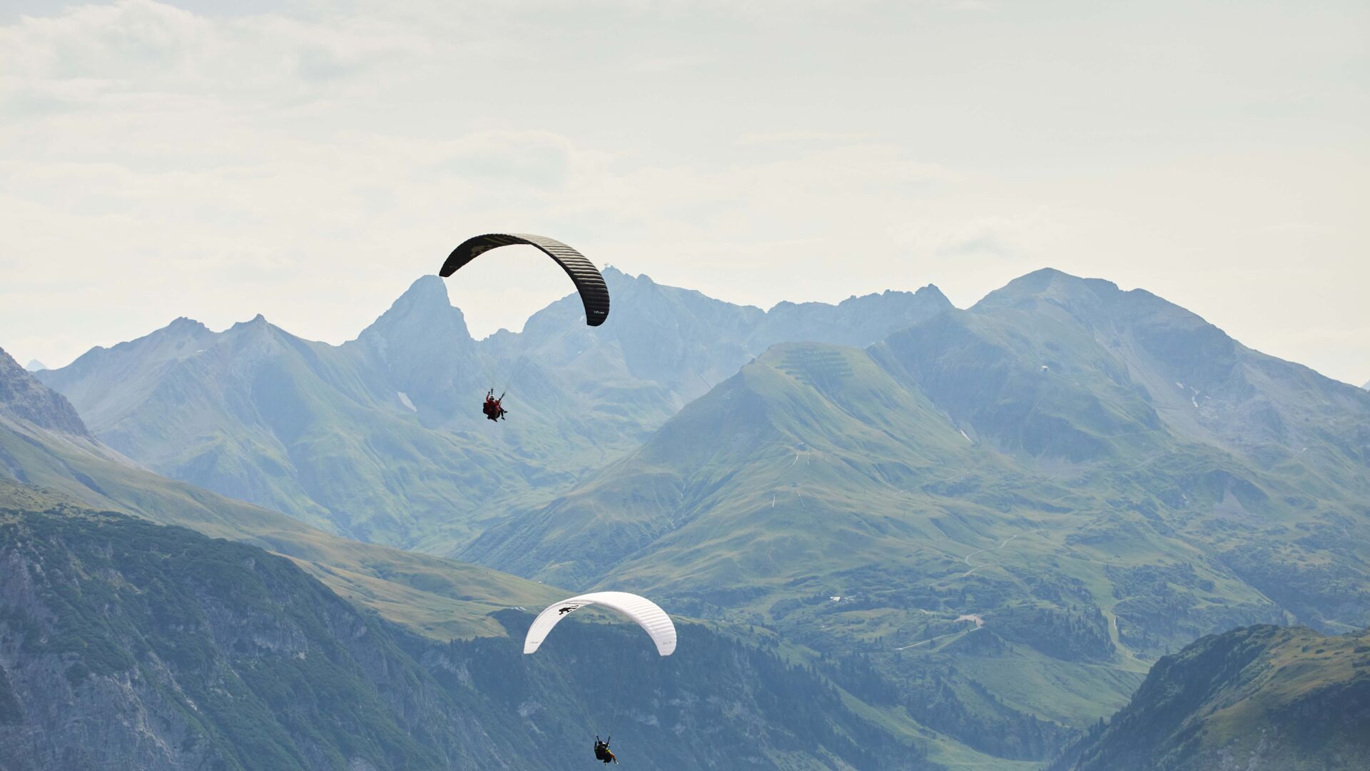 Zwei Paragleiter von hinten zu sehen und im Hintergrund eine große weite zu sehen mit Berggipfeln
