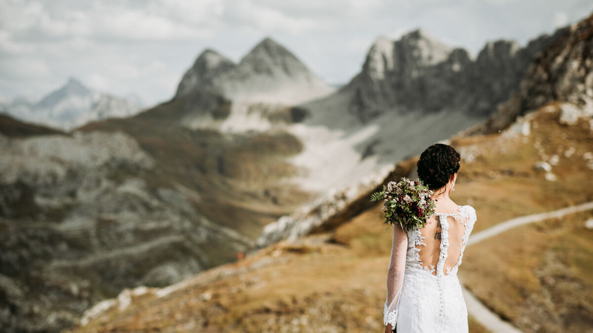 Braut steht von hinten sehend vor herrlicher Berglandschaft mit Blumenstrauß in der Hand