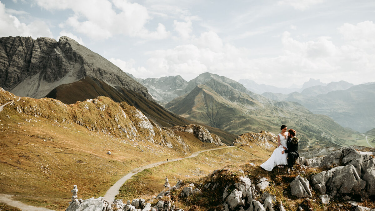 Frisch vermähltes Ehepaar sitzt auf Hügel vor traumhafter Berglandschaft und schaut sich in die Augen