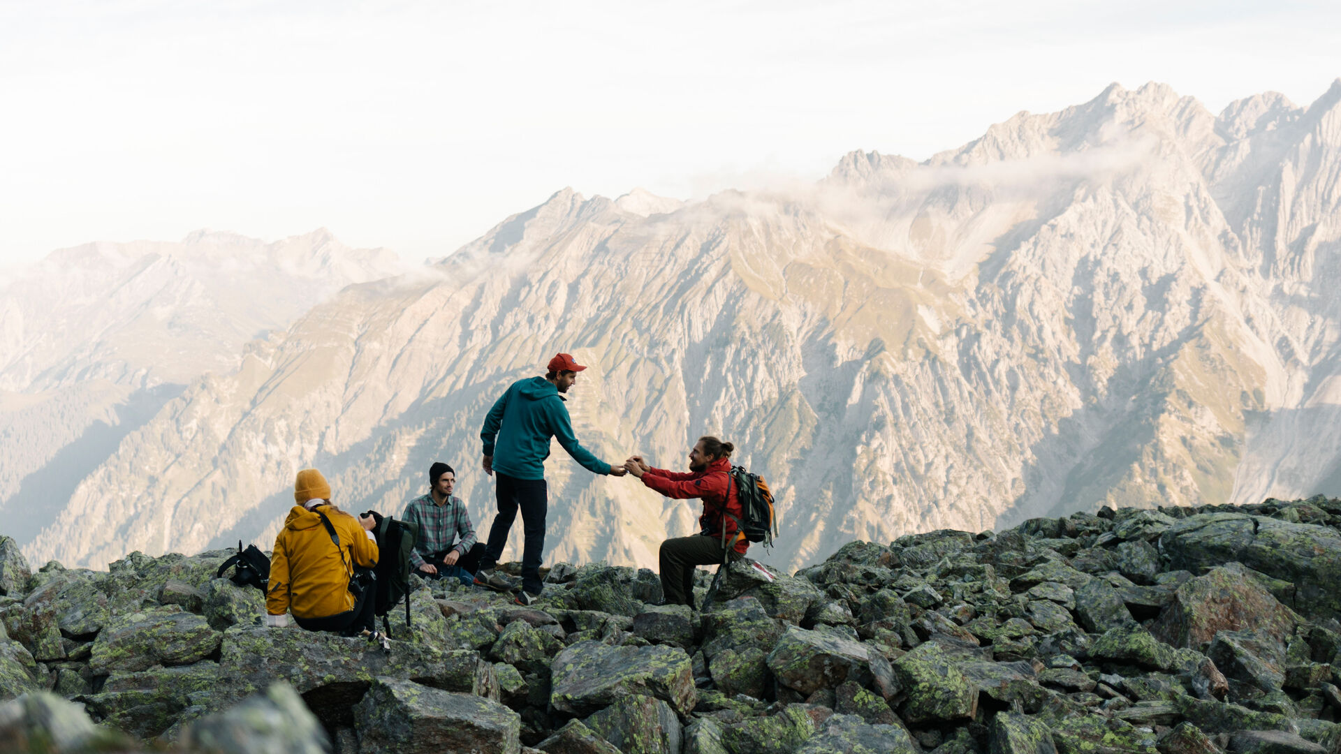 Vier Wanderer sitzen auf felsigem Boden. Im Hintergrund sieht man ein weitläufiges Gebirgspanorama. 