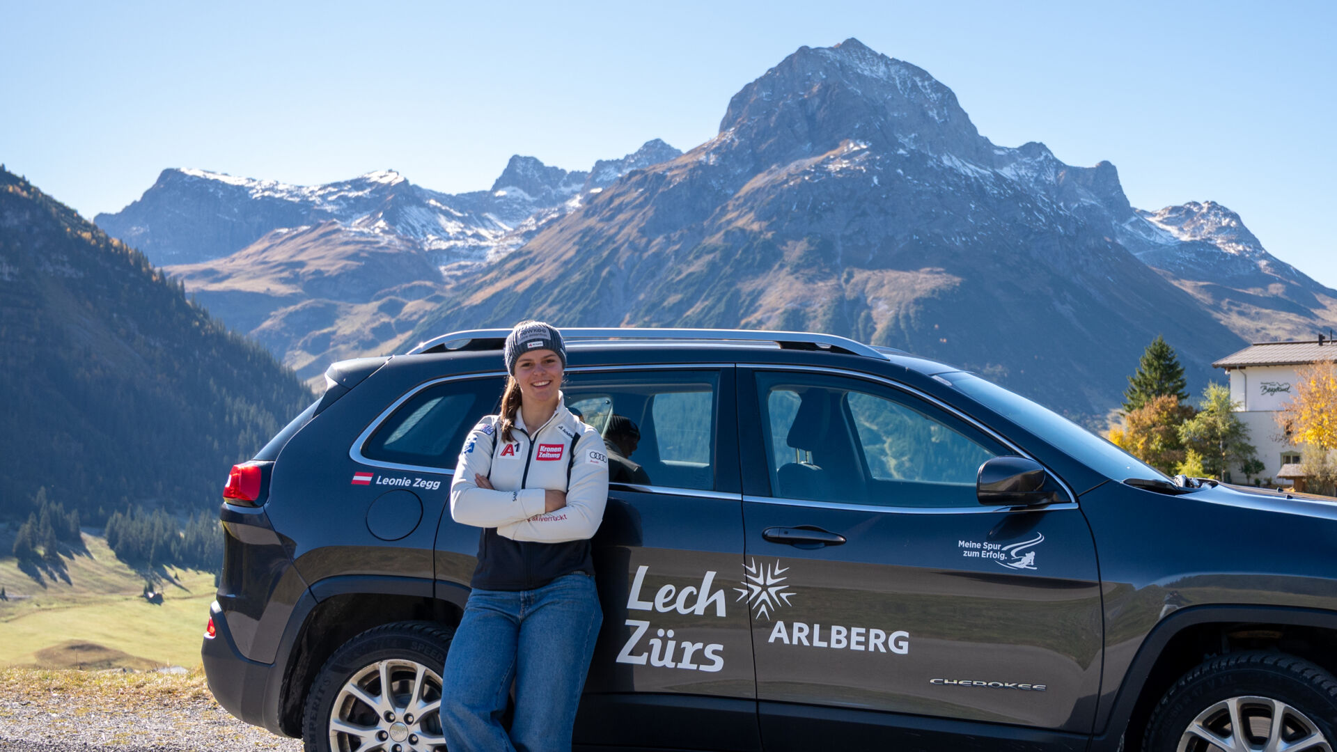 Frau steht vor Auto im Hintergrund ist das Omeshorn, der Hausberg von Lech zu sehen. Auf dem Auto ist ein großer Sticker-Aufdruck mit Lech Zürs Stern Arlberg zu sehen