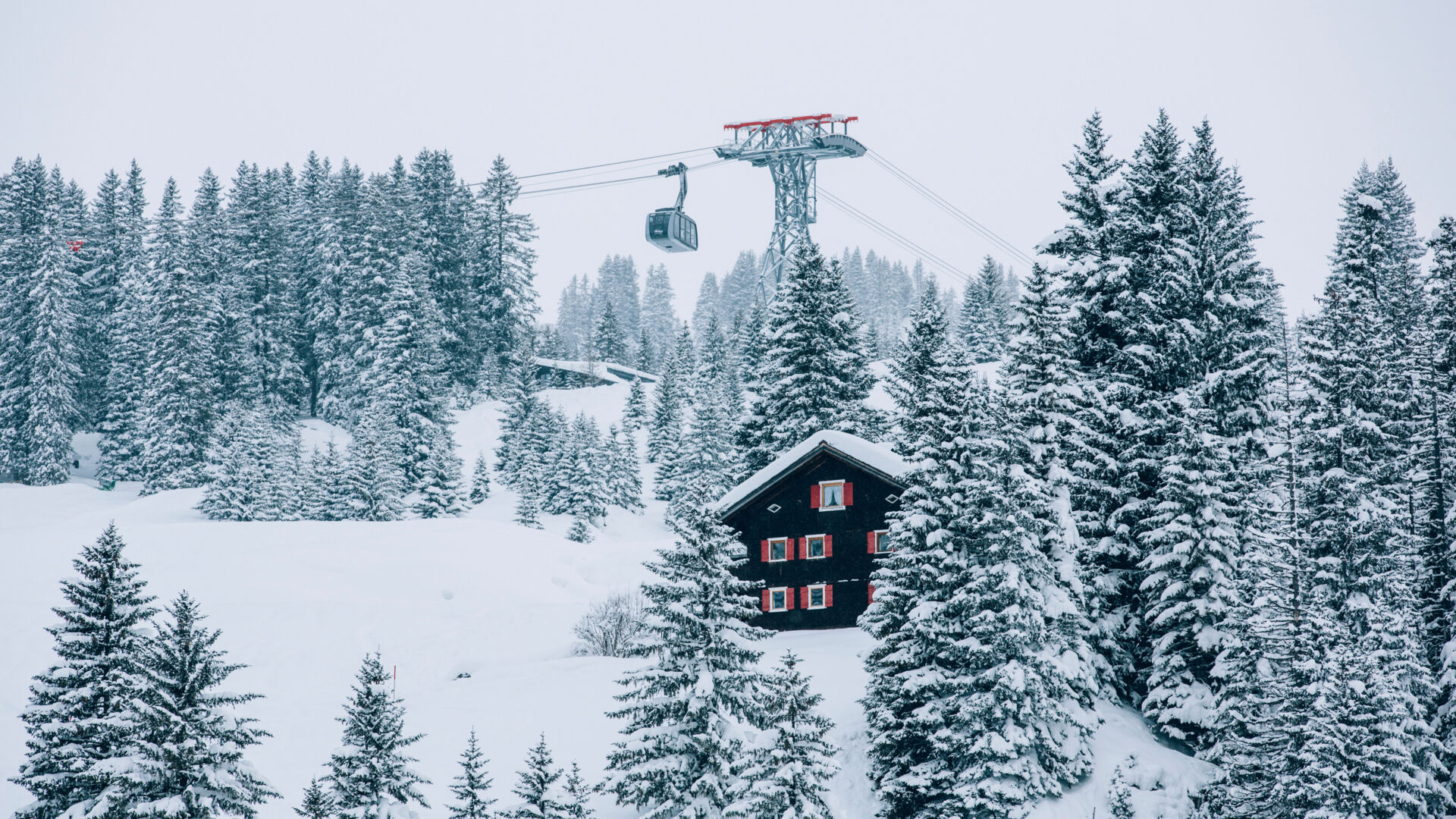 die Oberlecher Bergbahn fährt über die traumhafte Schneelandschaft, im Vordergrund ragt ein uriges Holzhaus zwischen den verschneiten Tannenbäumen heraus