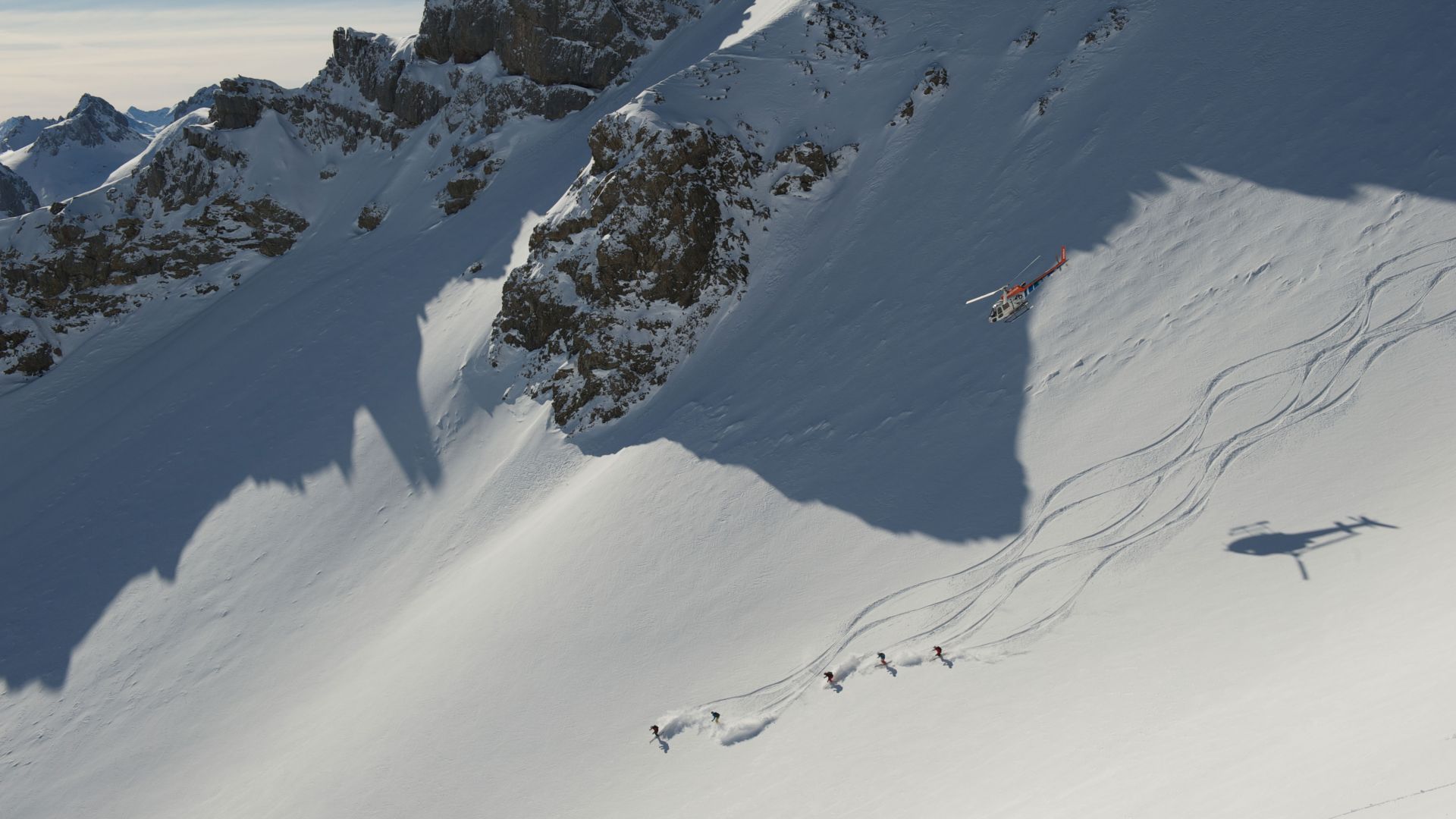 Blick auf eine Gruppe beim Heliskiing, die die ersten Schwünge in der unbefahrenen Offpiste fahren, über ihnen fliegt der Helikopter