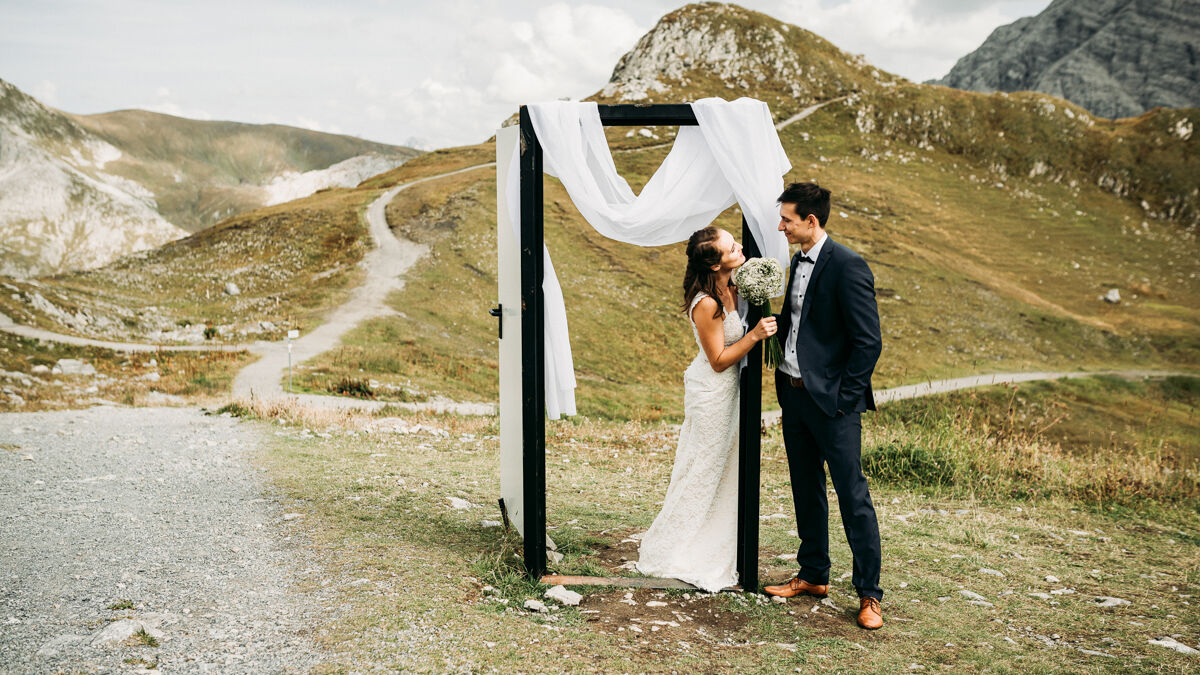 Brautpaar steht vor Bergkulisse und schaut sich in die Augen. Im Hintergrund ist ein kleiner Schleier über einen Traubogen gelegt.