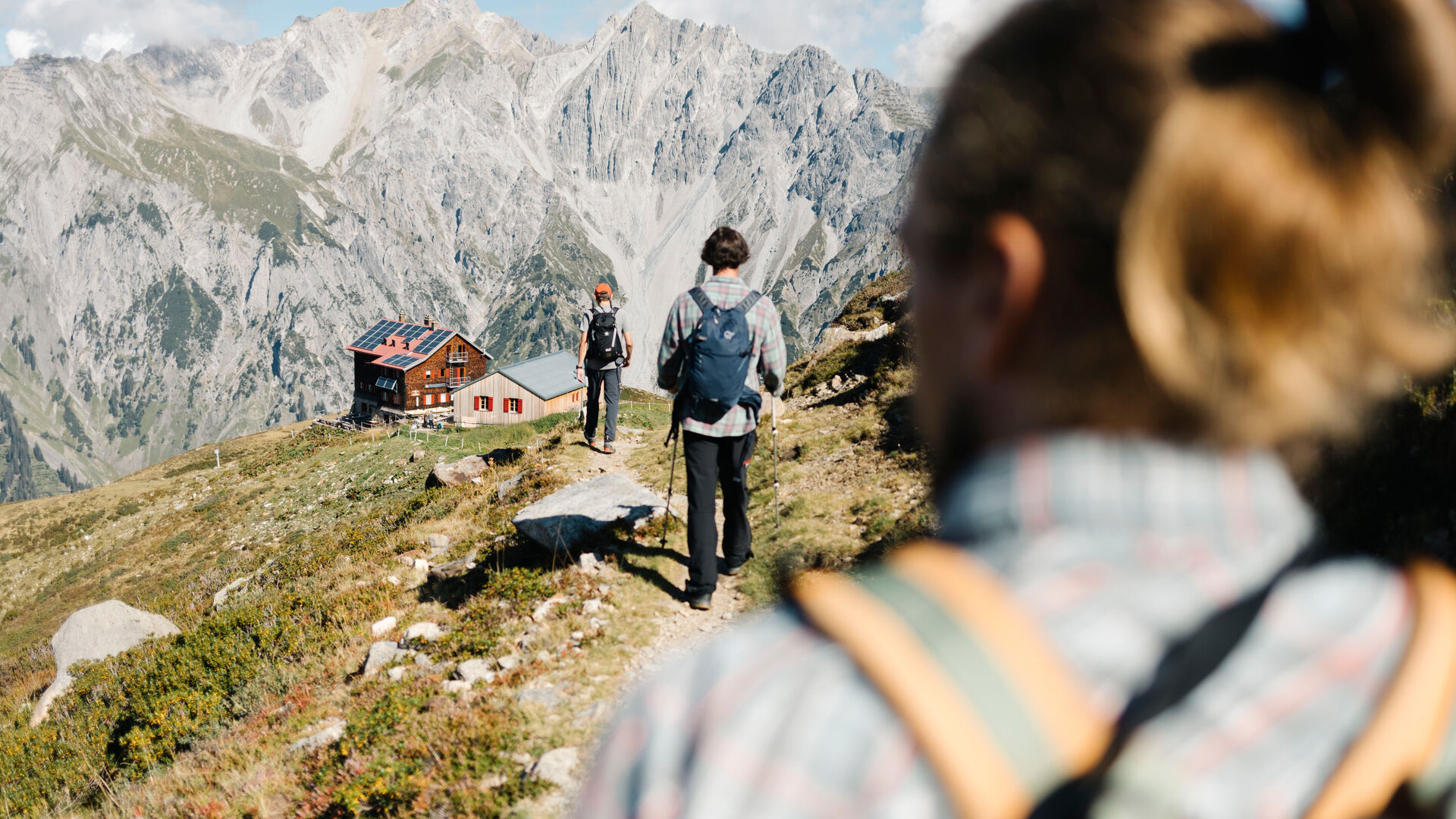 Drei Wanderer gehen in die Richtung einer Berghütte. Im Hintergrund ist ein felsiges Gebirgspanorama.