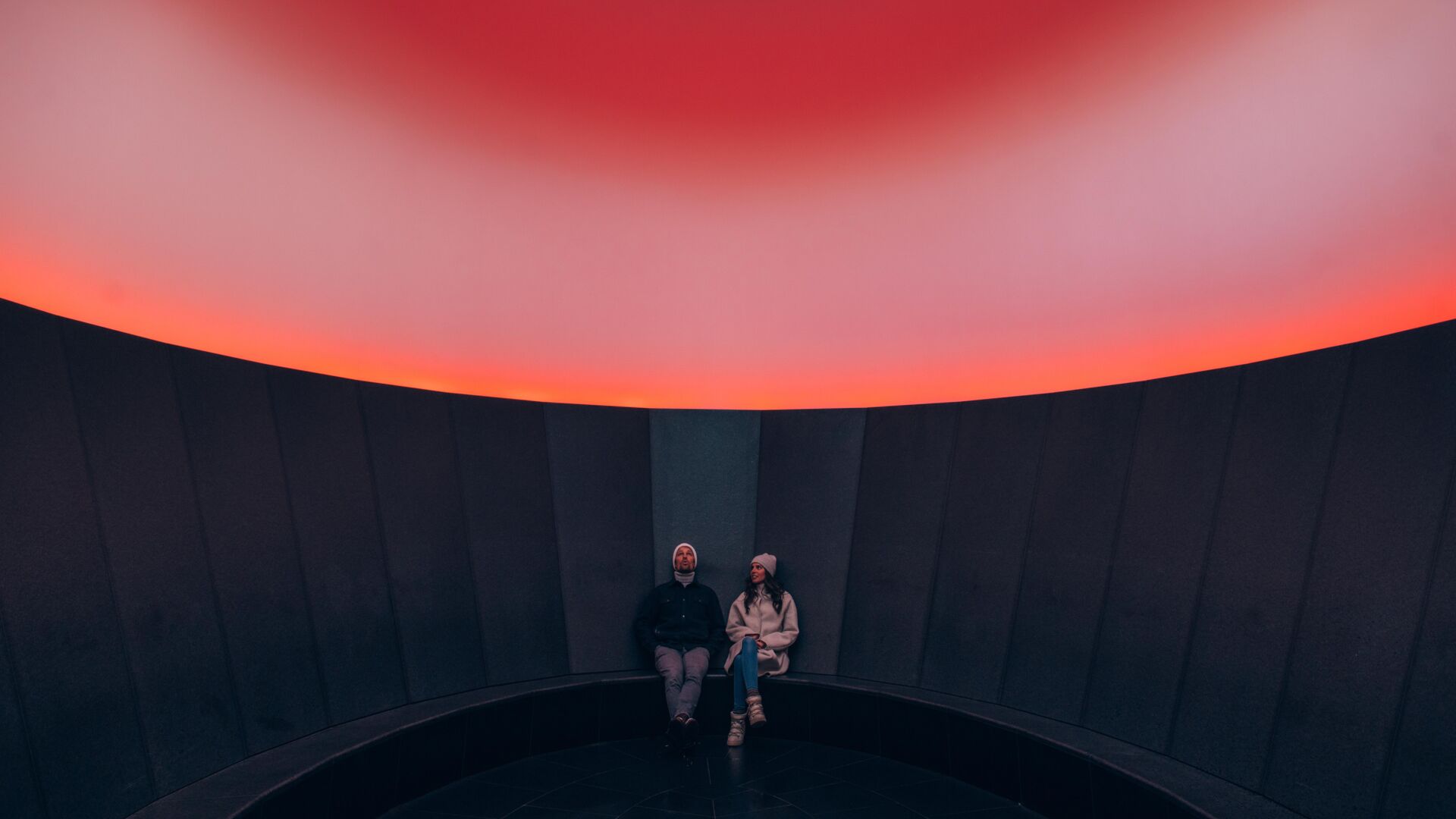 zwei Personen betrachten das Lichtspektakel im Skyspace, durch die Sonneneinstrahlung erscheint es in einem rötlichen Farbspiel