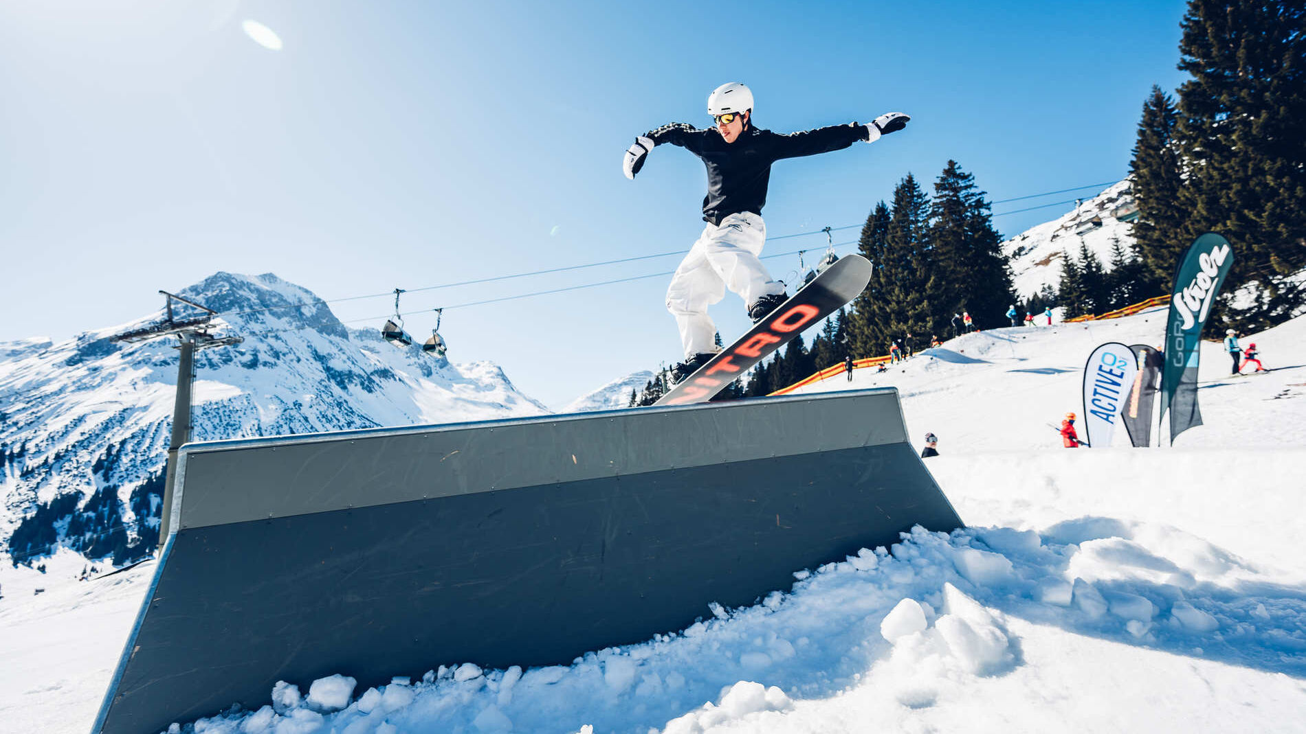 Freestyler fährt über eine Rail und im Hintergrund ist strahlend blauer Himmel und eine wunderschöne Bergkulisse zu sehen.